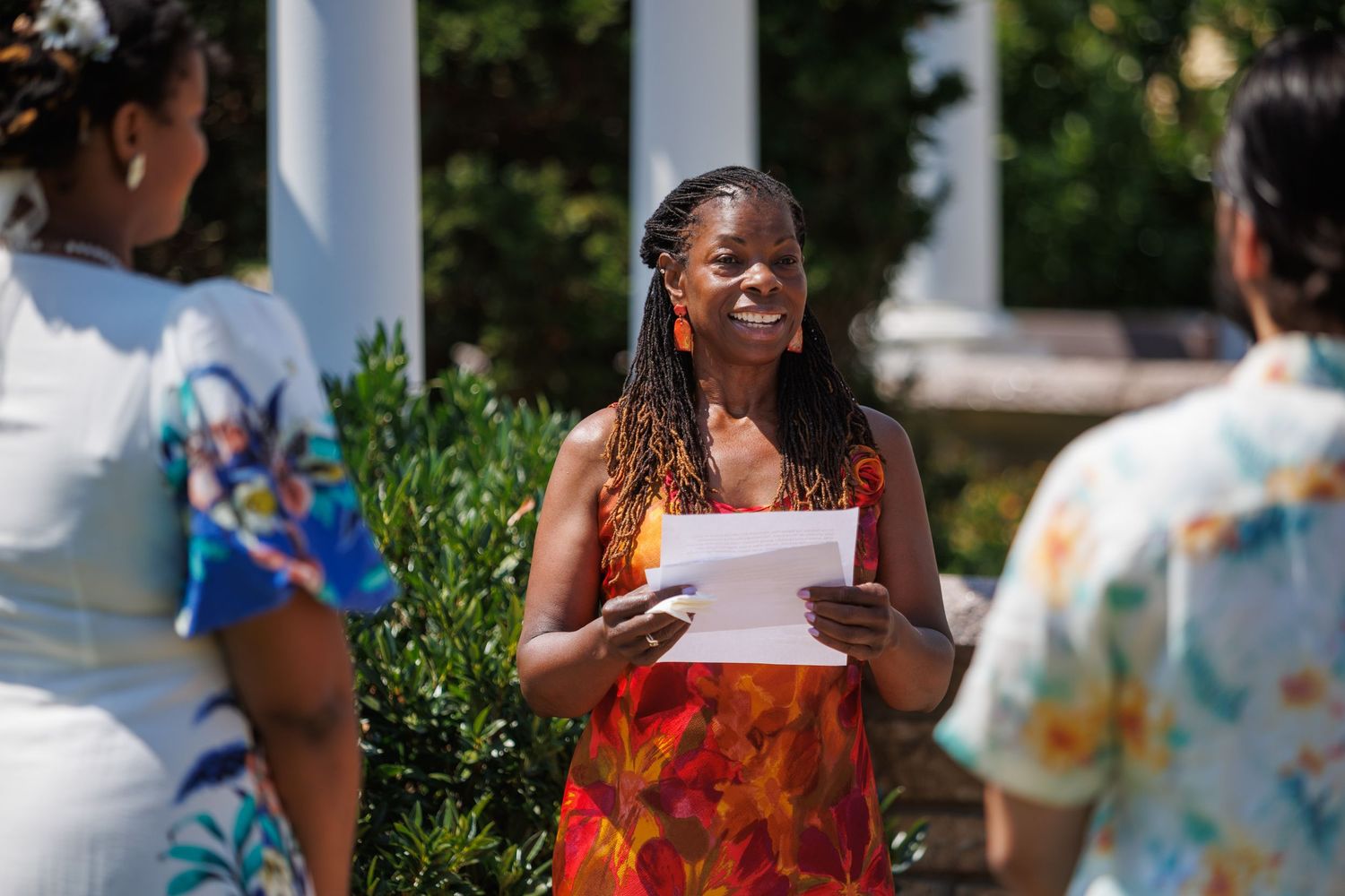 A smiling Black woman in a colorful dress holds papers outdoors at a sunny garden ceremony with white columns.