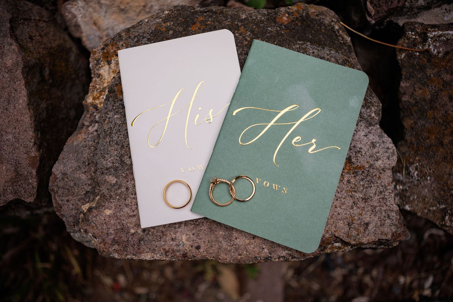 His and Hers vow books in white and sage green with gold lettering and wedding bands on stone.
