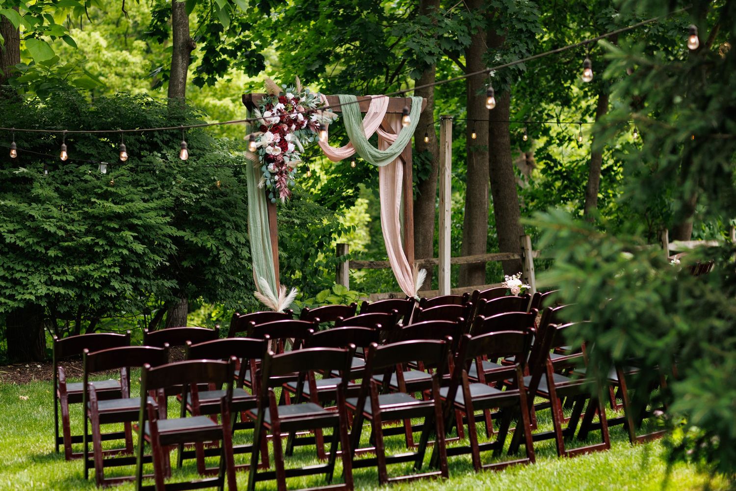 Outdoor wedding ceremony setup with dark wooden chairs, floral arch, and string lights among lush green trees.