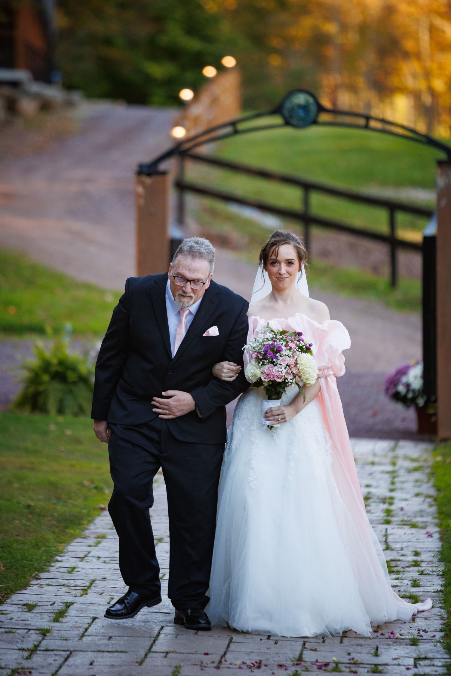 Father escorts smiling bride in white tulle gown with pink accents down garden pathway at sunset wedding.