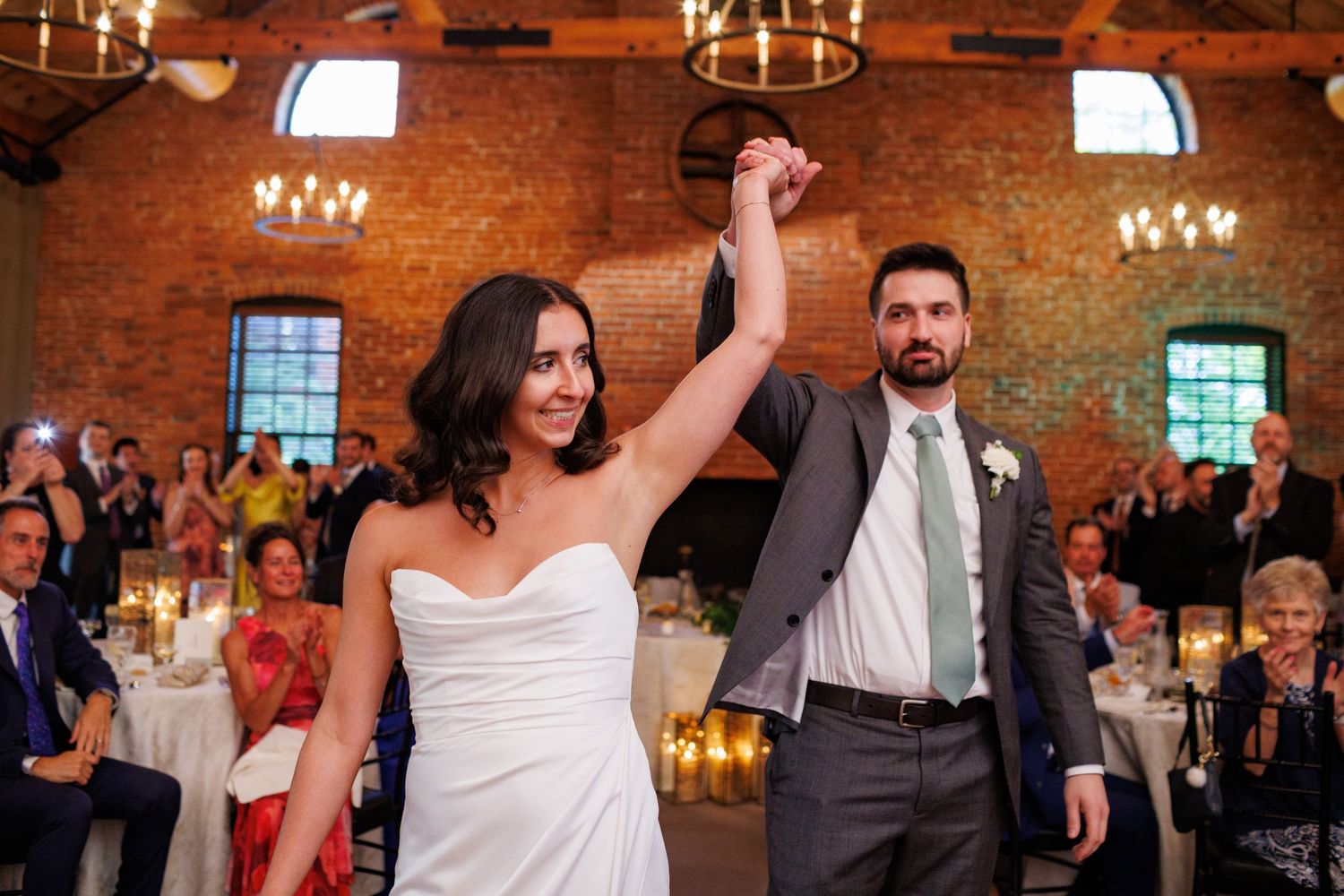 Bride and groom make grand entrance into rustic brick reception hall as guests applaud and celebrate.