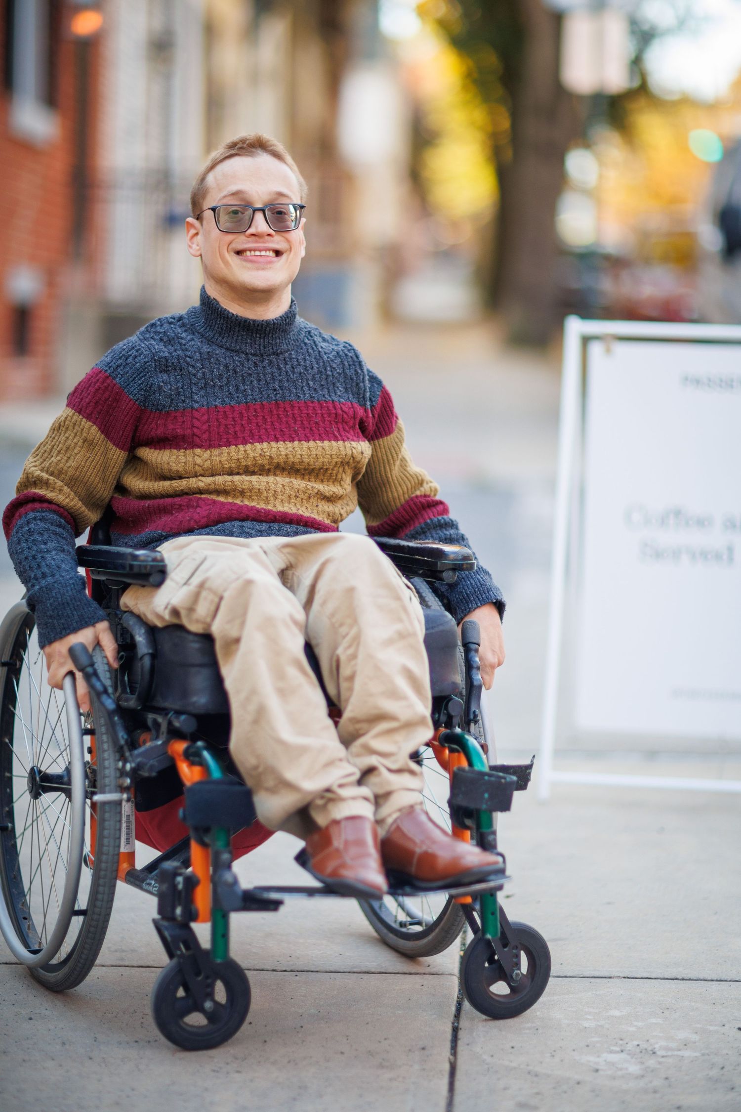 A person in a striped sweater sits in a wheelchair on a city sidewalk in autumn.