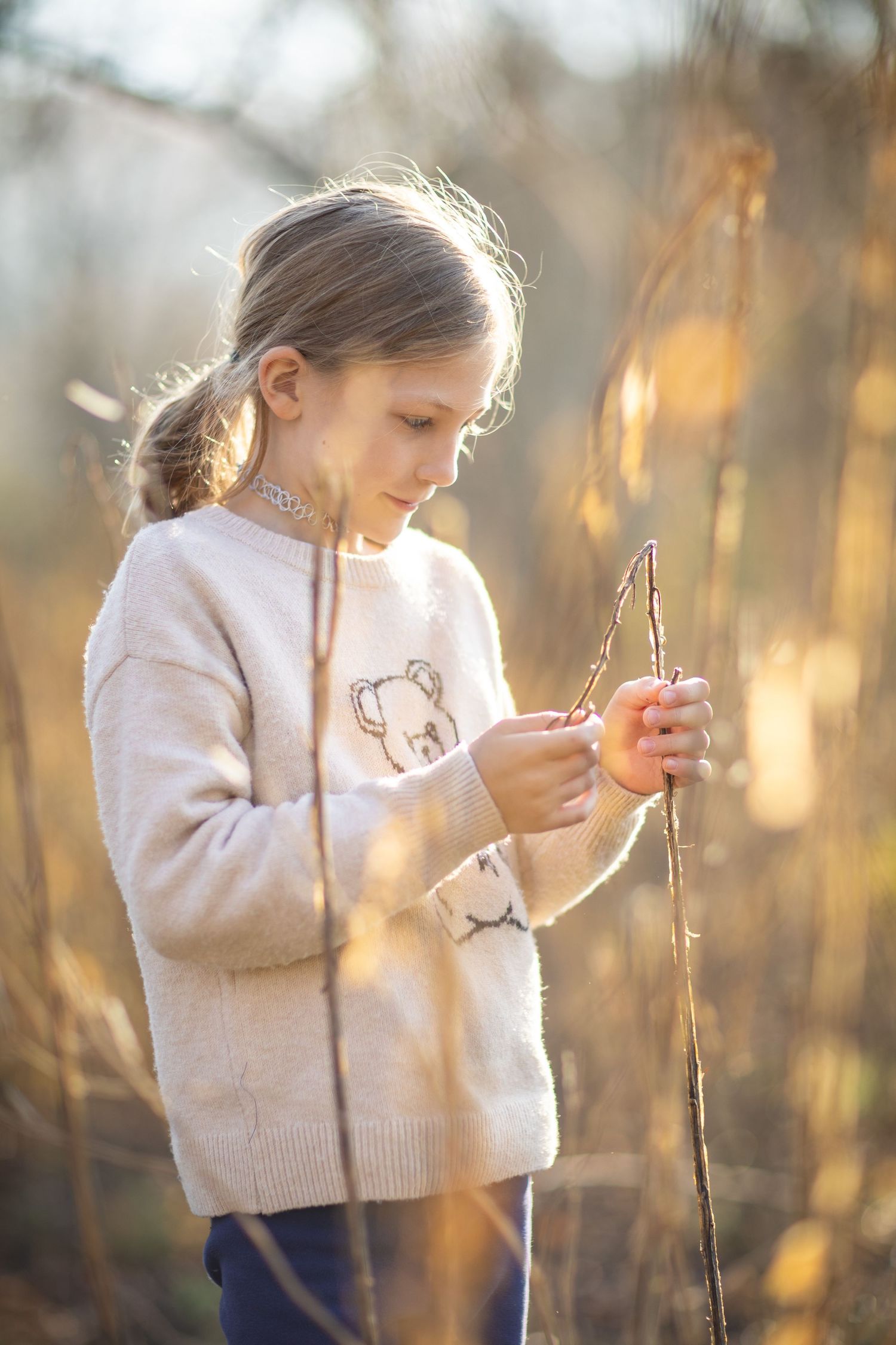 A young child in a cream sweater examines a tree branch during golden hour in nature.