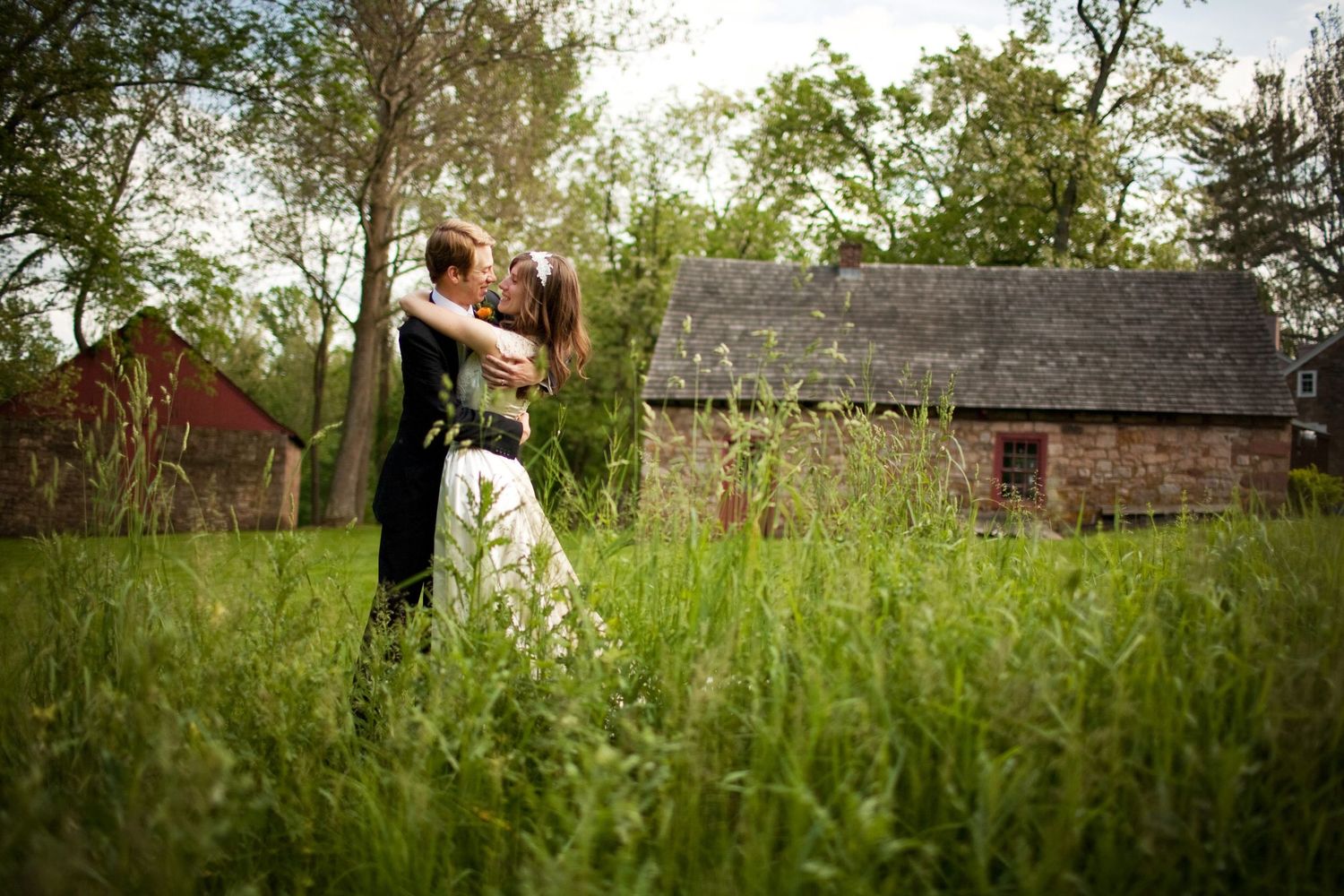 A romantic couple embraces in tall grass with rustic barns in the background during a countryside wedding photoshoot.