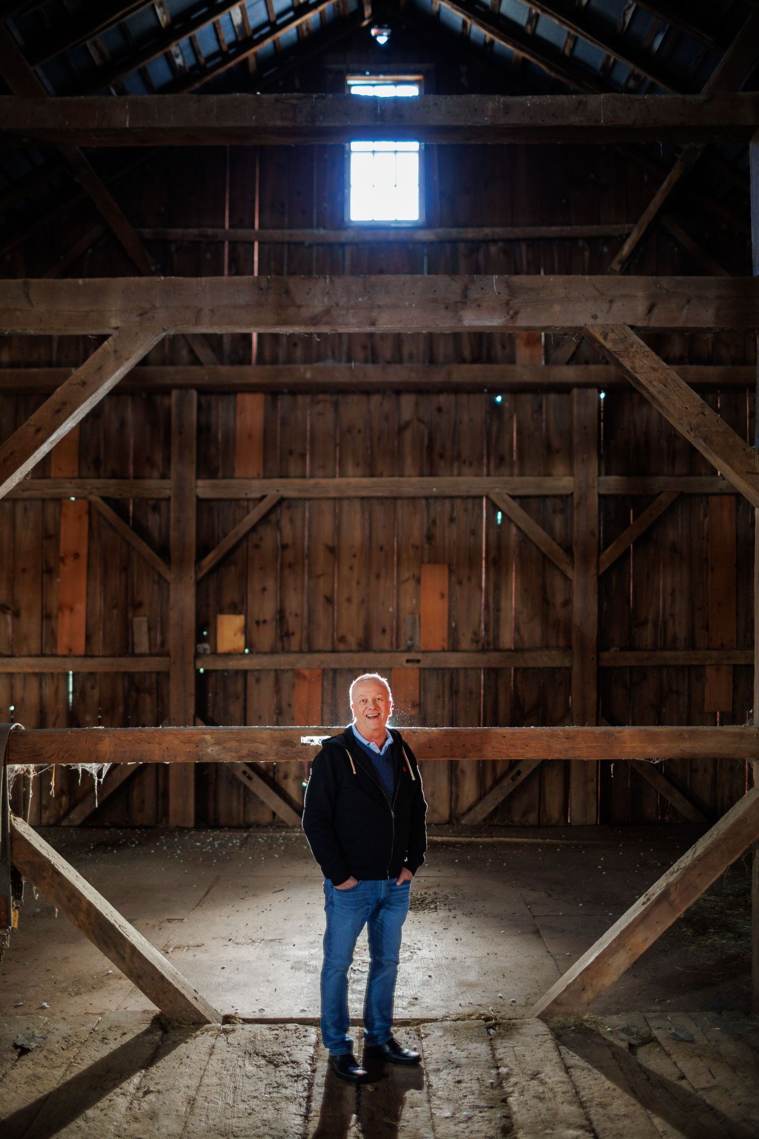 A rustic wooden barn interior with sunlight streaming through gaps in the walls illuminates a figure sitting on a beam.