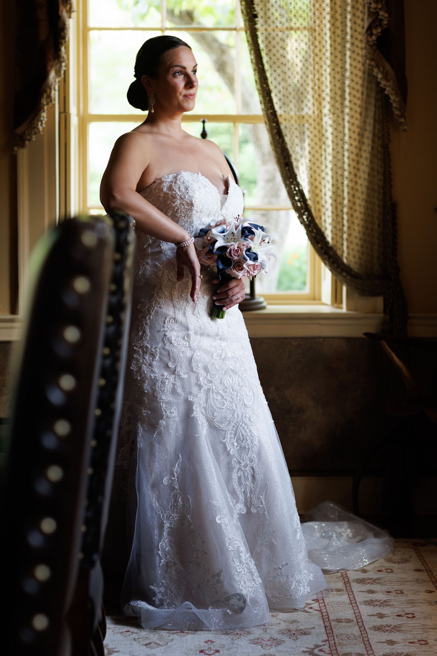 A bride in a white lace wedding gown stands by a window in a vintage-style room with natural lighting.