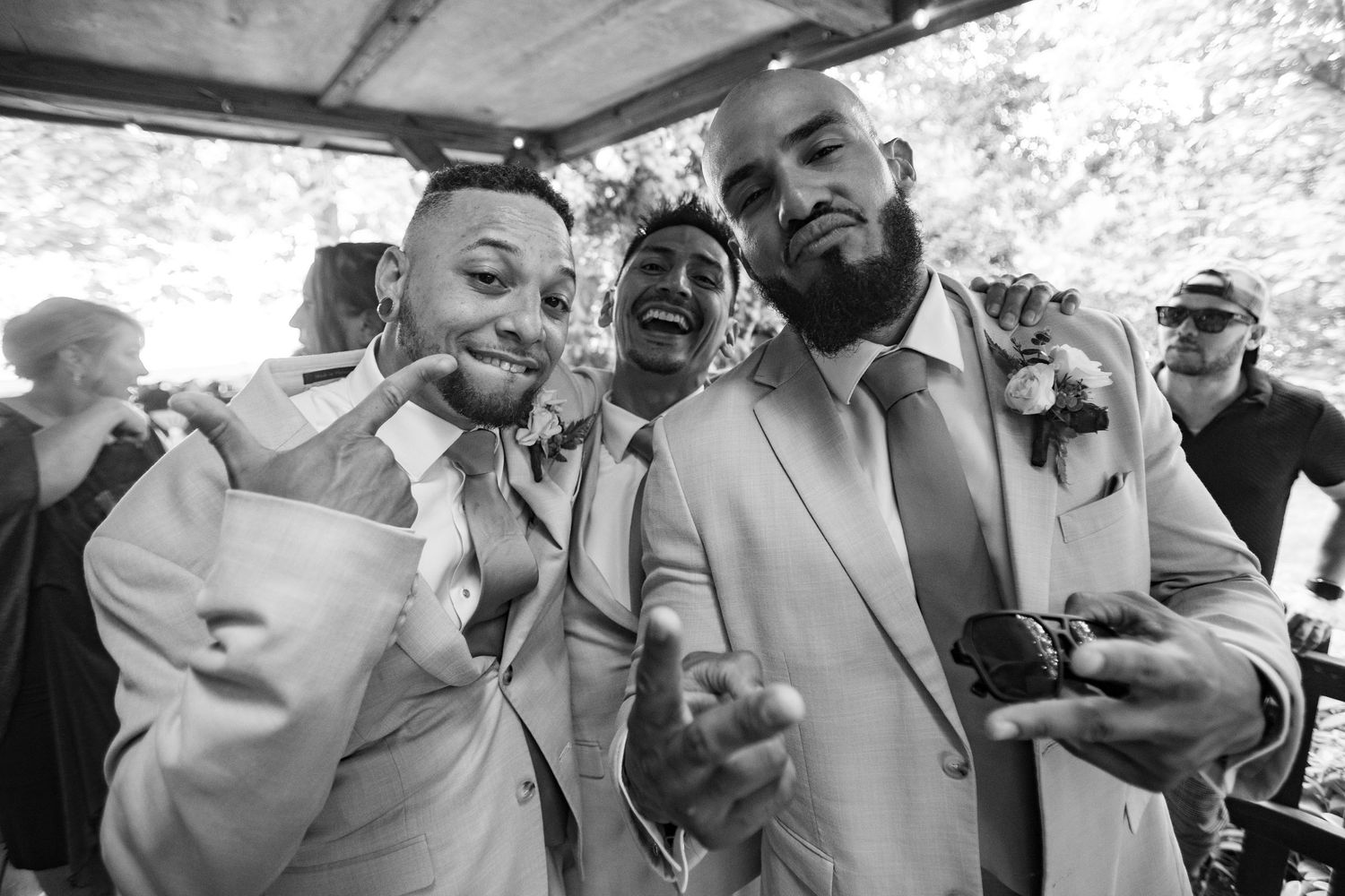 Three groomsmen in tan suits laugh and pose together at outdoor wedding reception.
