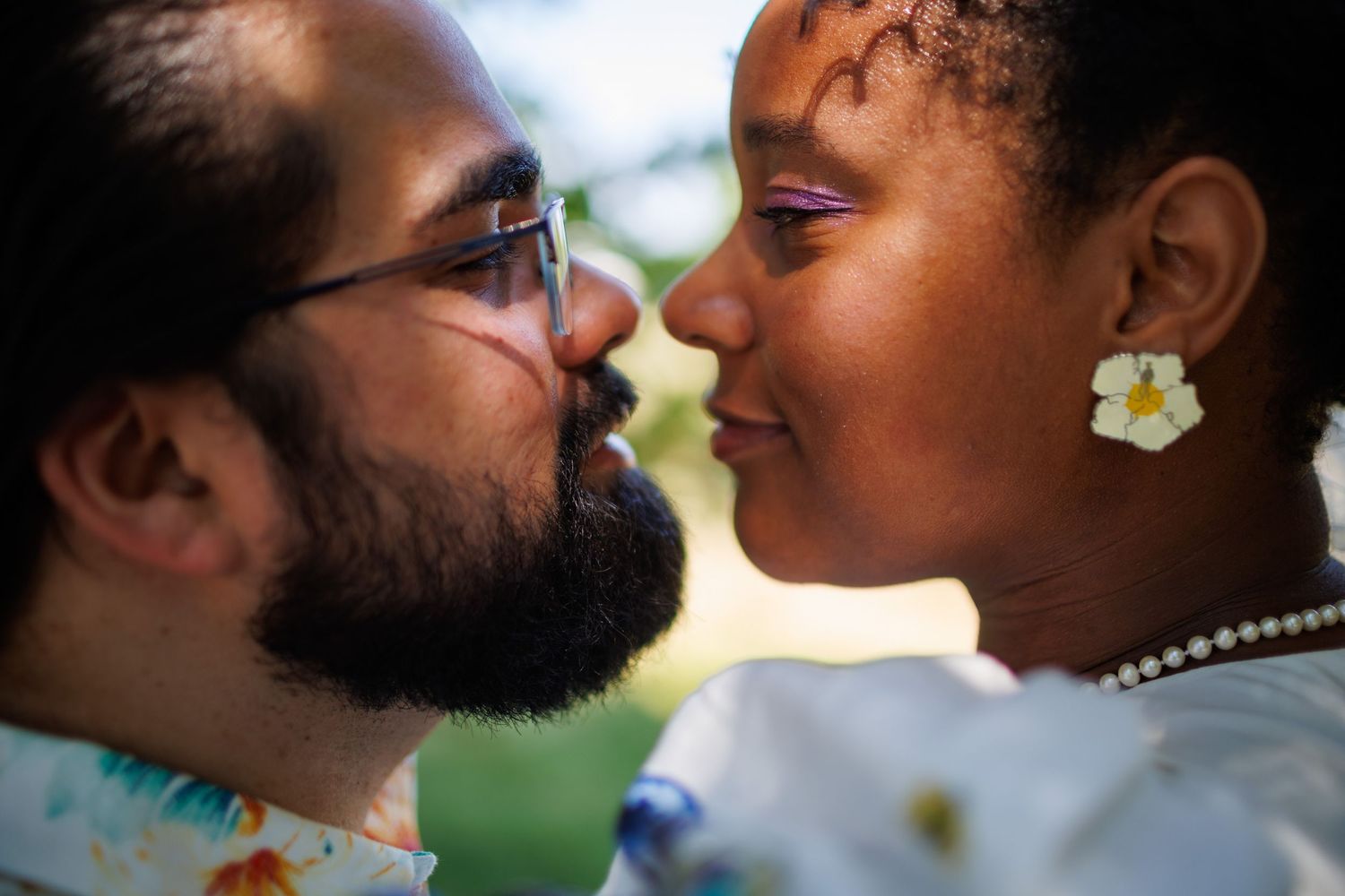 Interracial couple sharing a romantic moment, woman wearing pearl necklace and white flower earring, man with beard and glasses.