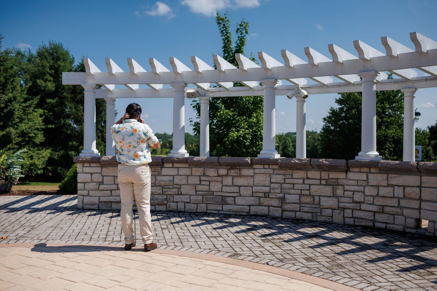 A man in a floral shirt stands before a white pergola with stone columns and brick pavers in a sunny garden.