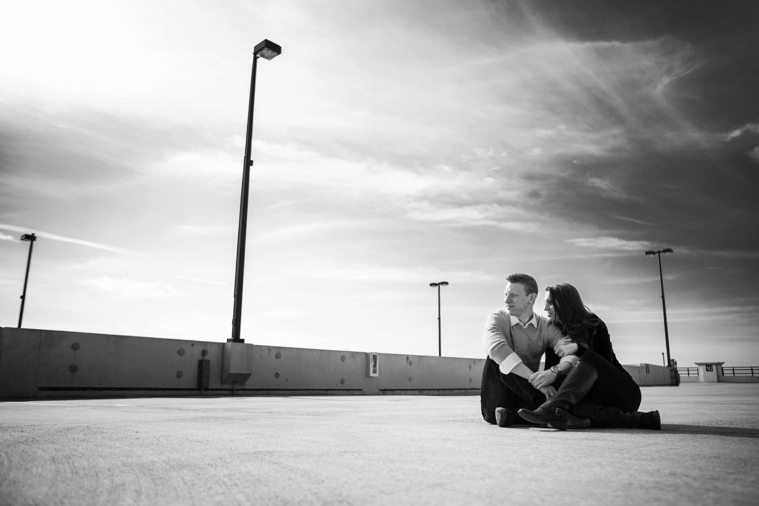 Black and white photo of a couple sitting intimately together on a rooftop under lampposts and cloudy skies.