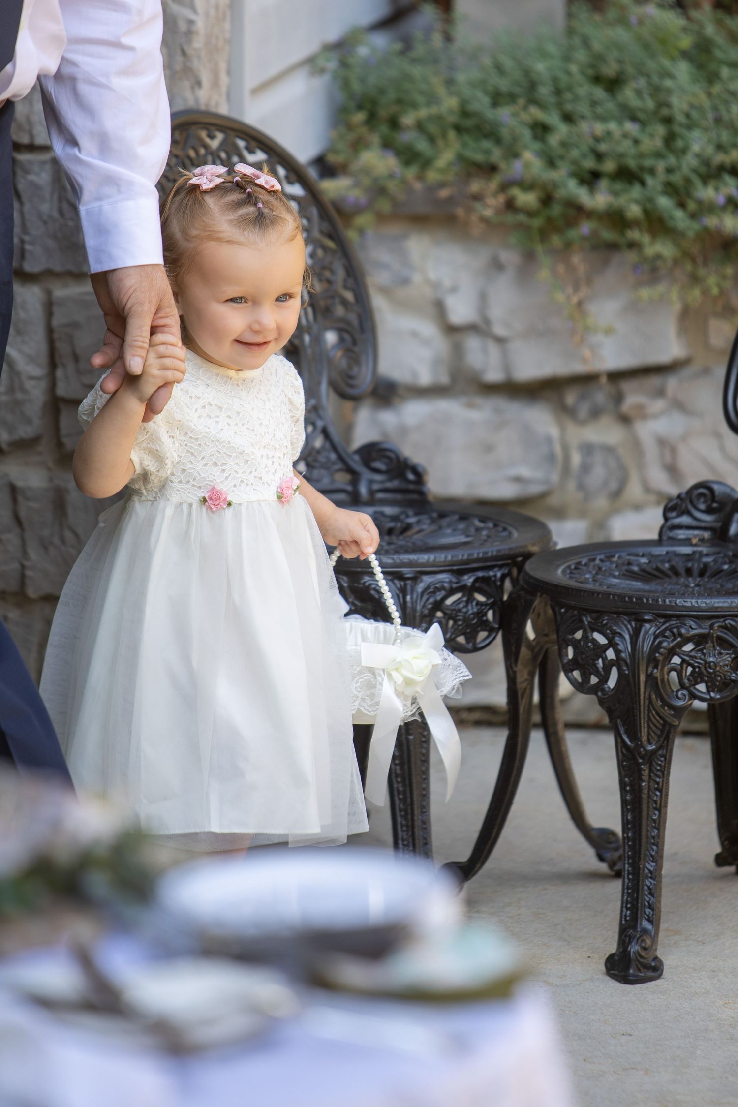 Small child in white dress holding adult's hand while walking beside ornate black metal bench in outdoor setting.