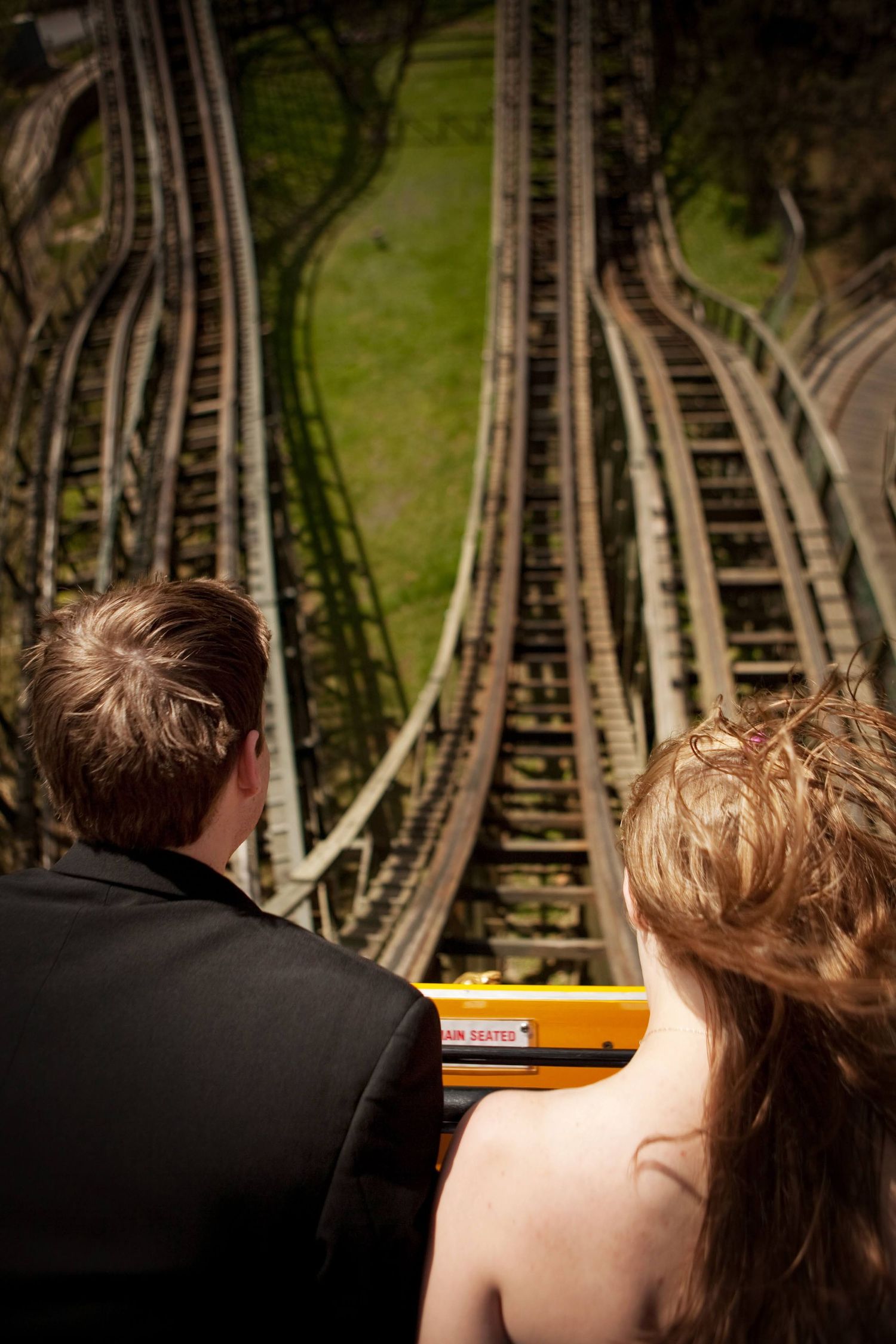 Two people sit in front view of a roller coaster track, with green grass and winding coaster rails visible ahead.