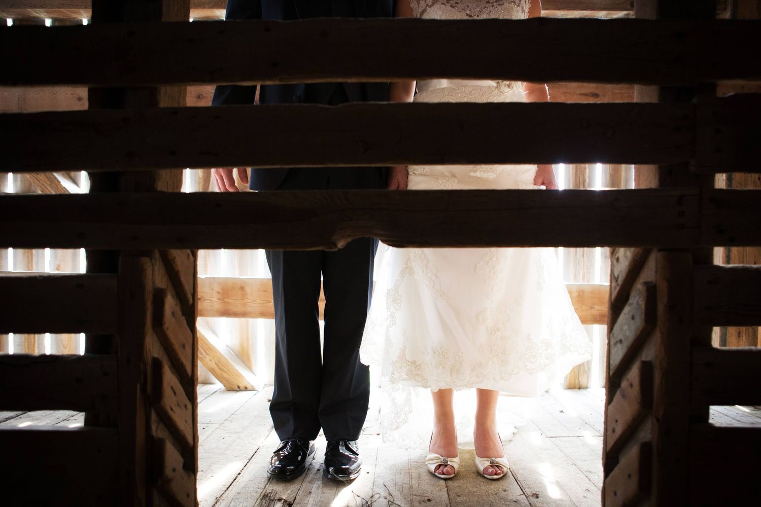 View through wooden slats showing feet standing on a rustic floor, with dark pants and a white dress visible.