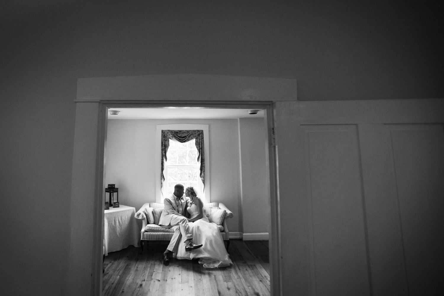 Black and white image of a couple in a bright window-lit room with a wedding dress hanging above.