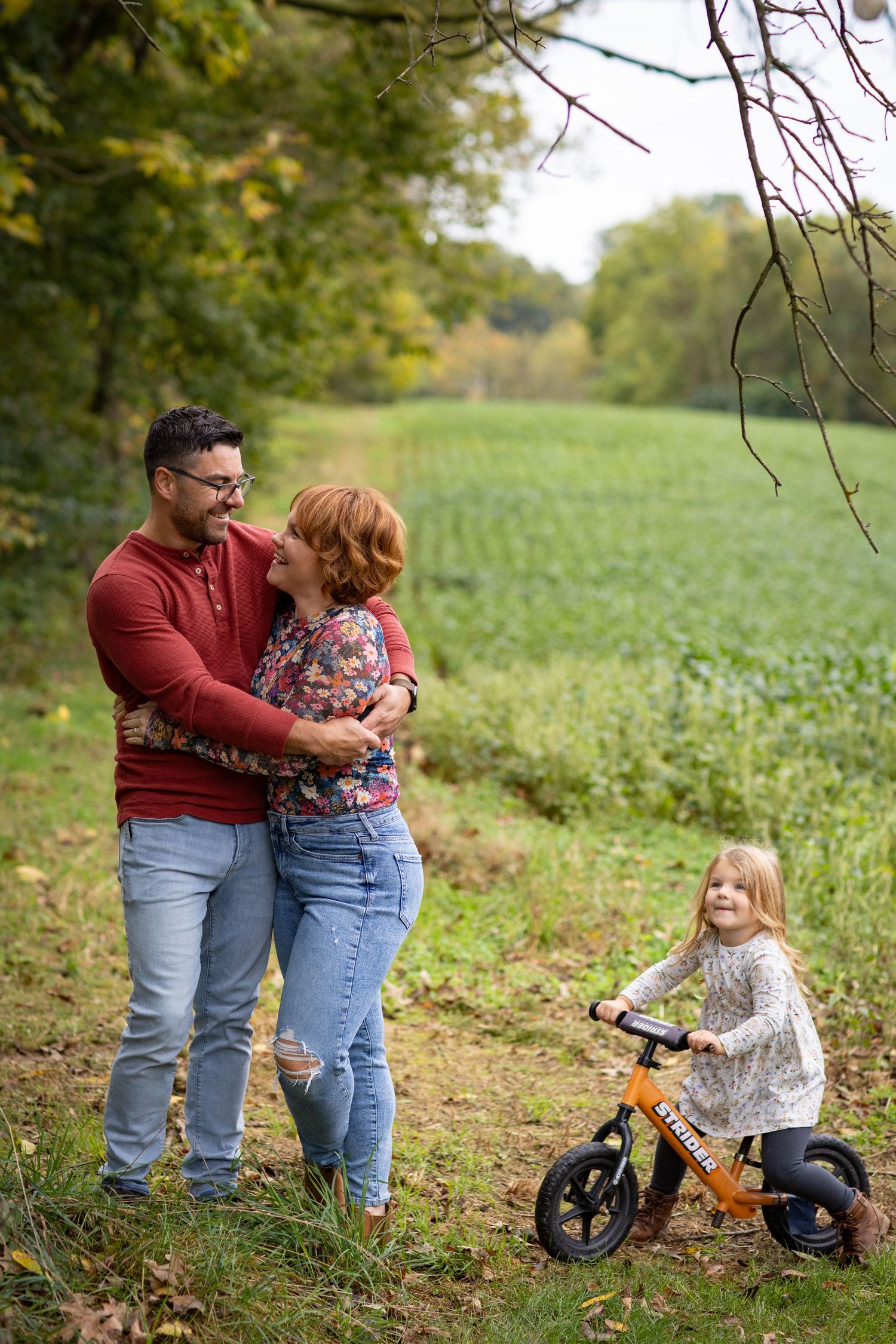 A couple and child enjoy a fall day outdoors with a balance bike in a green field surrounded by trees.