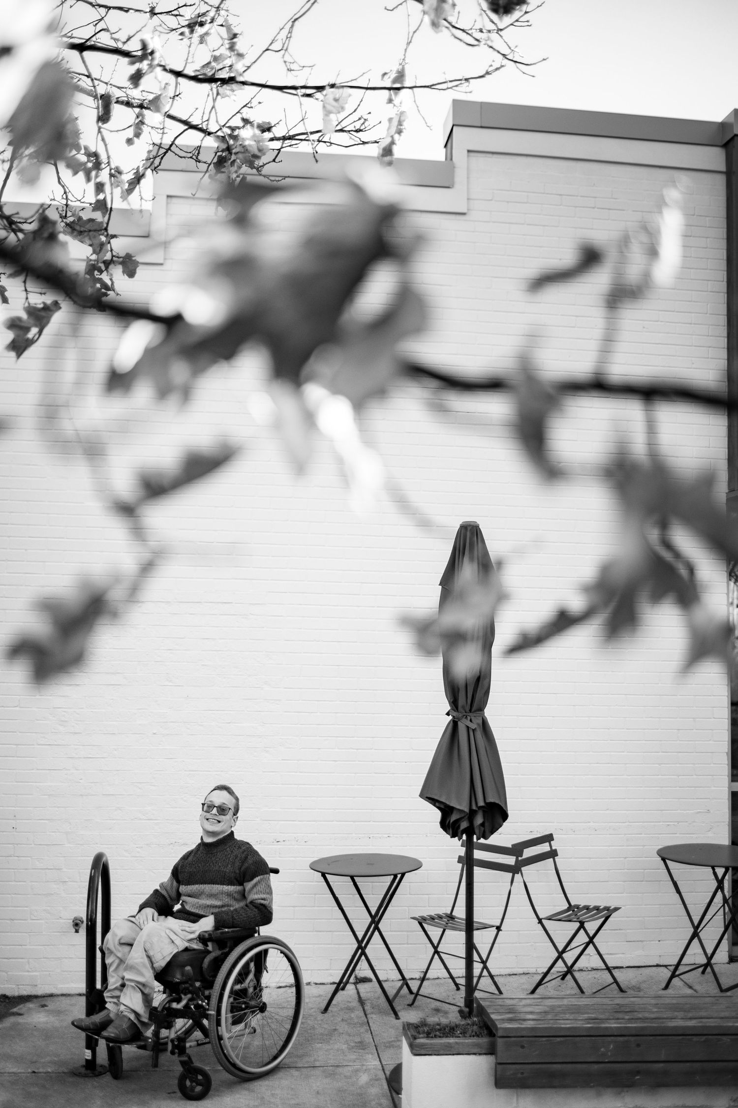 Black and white photo of person in wheelchair sitting on outdoor patio with cafe chairs and umbrella against white wall.