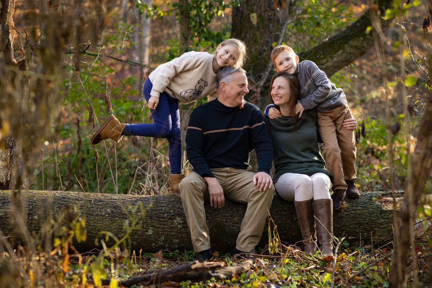 A family of four sits together on a rustic wooden bench surrounded by autumn foliage in a natural outdoor setting.