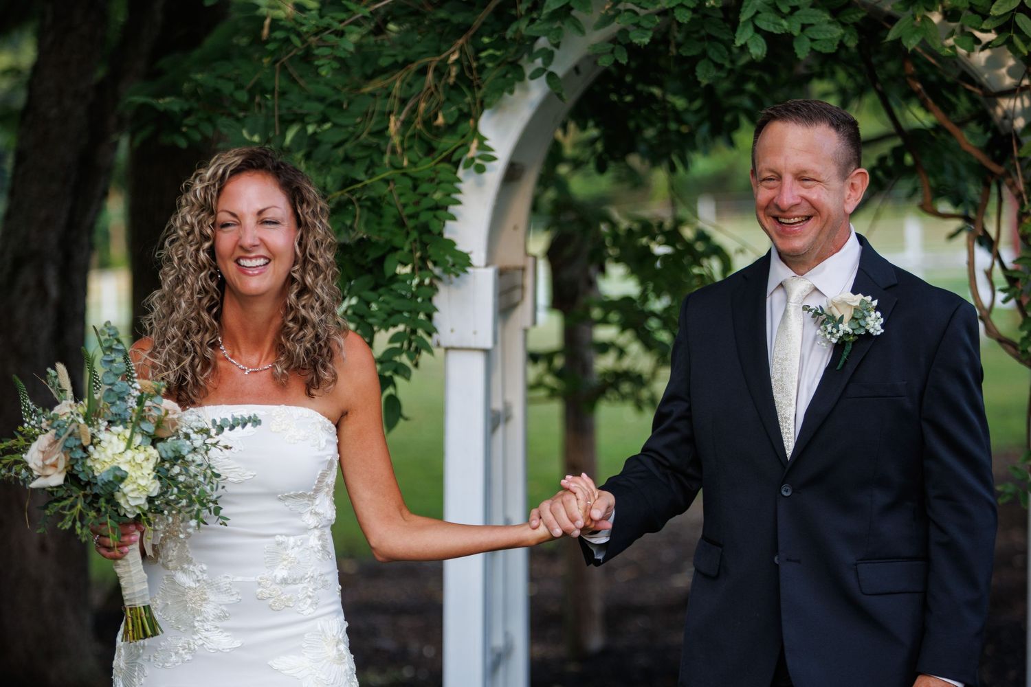 Smiling bride and groom hold hands under a white arbor surrounded by lush green foliage.