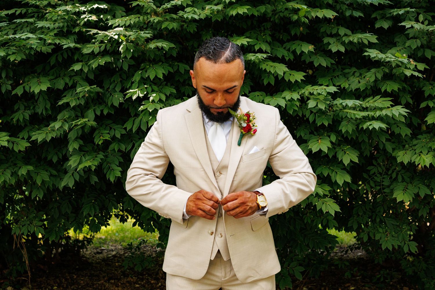 Handsome groom in a beige suit adjusting his jacket buttons in front of a lush green hedge.