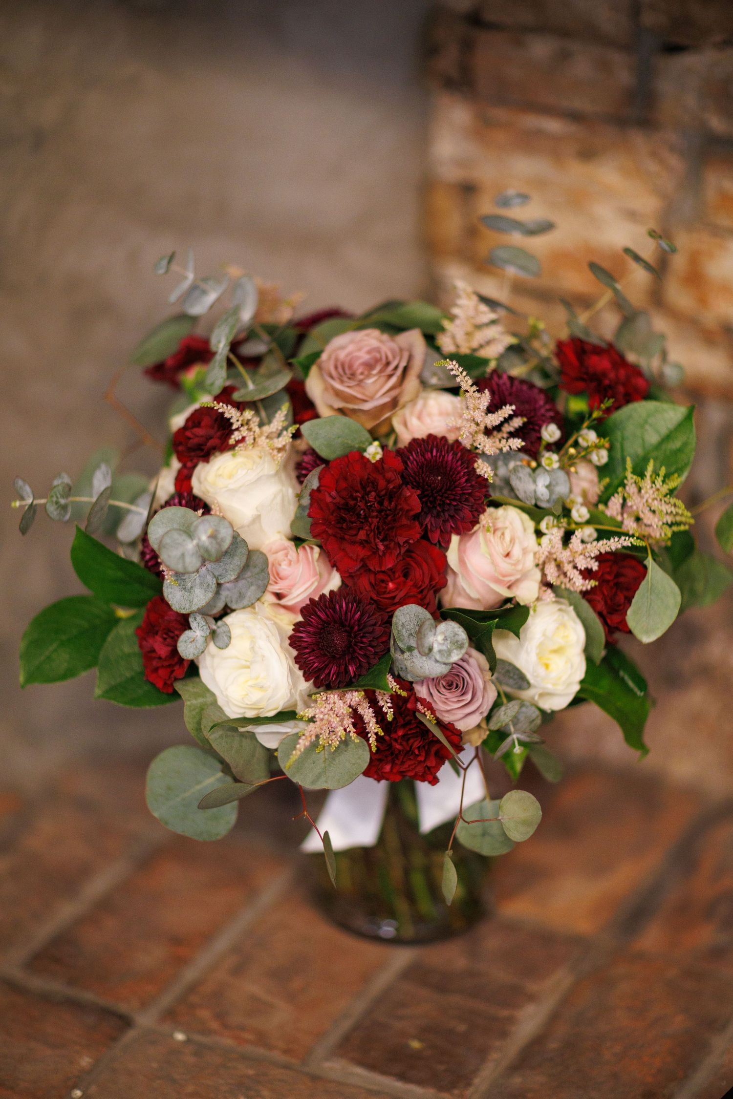 Elegant bridal bouquet featuring deep red dahlias, blush roses, white flowers, and eucalyptus greenery in a glass vase.