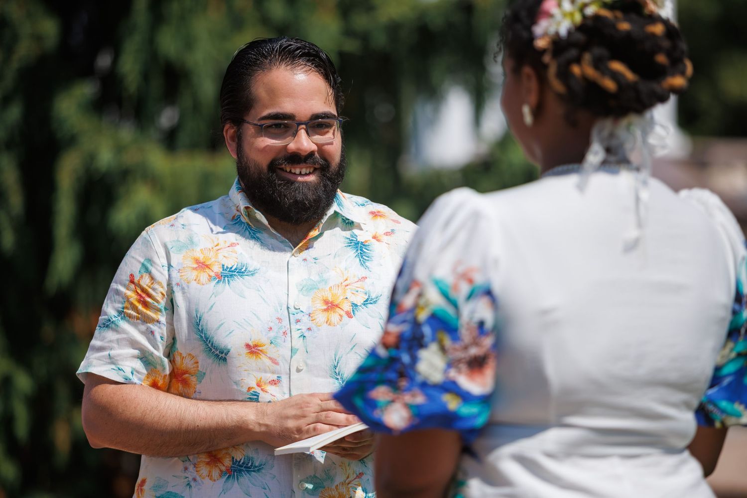Smiling bearded man in floral shirt during outdoor wedding ceremony on sunny day.