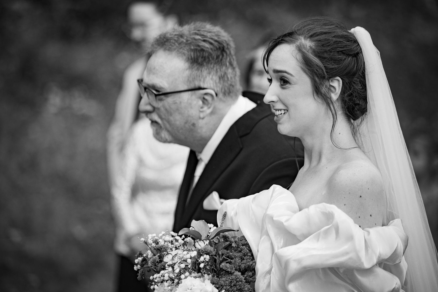 Black and white photo of a smiling bride in a white off-shoulder gown and veil walking with an older man at a wedding.