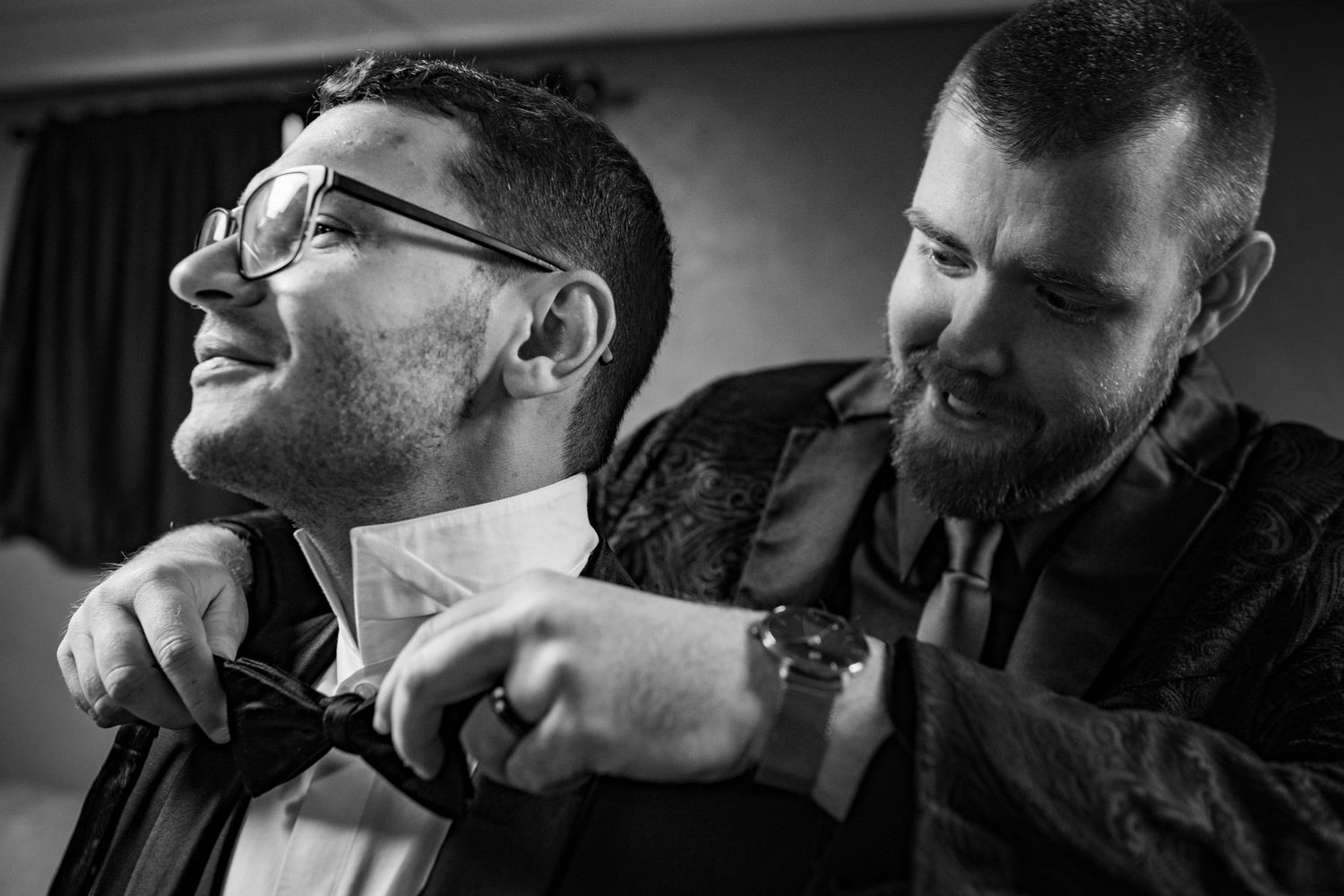 Groom smiling as best man adjusts his bow tie in black and white wedding preparation photo.