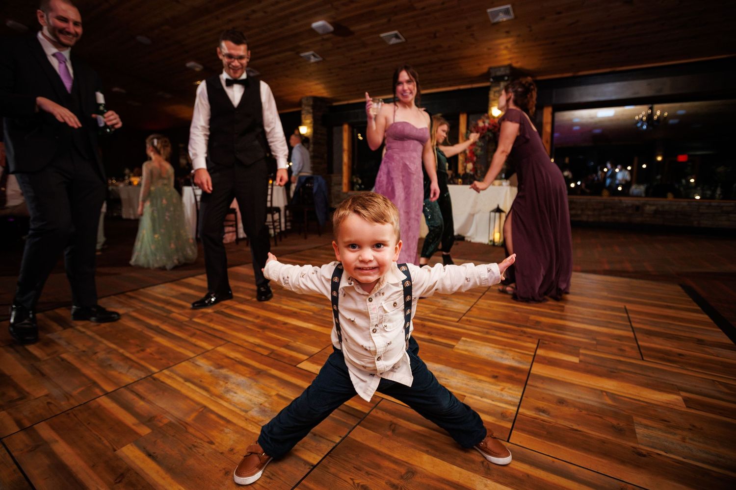 Excited young boy strikes a wide stance pose on the wedding reception dance floor surrounded by guests.