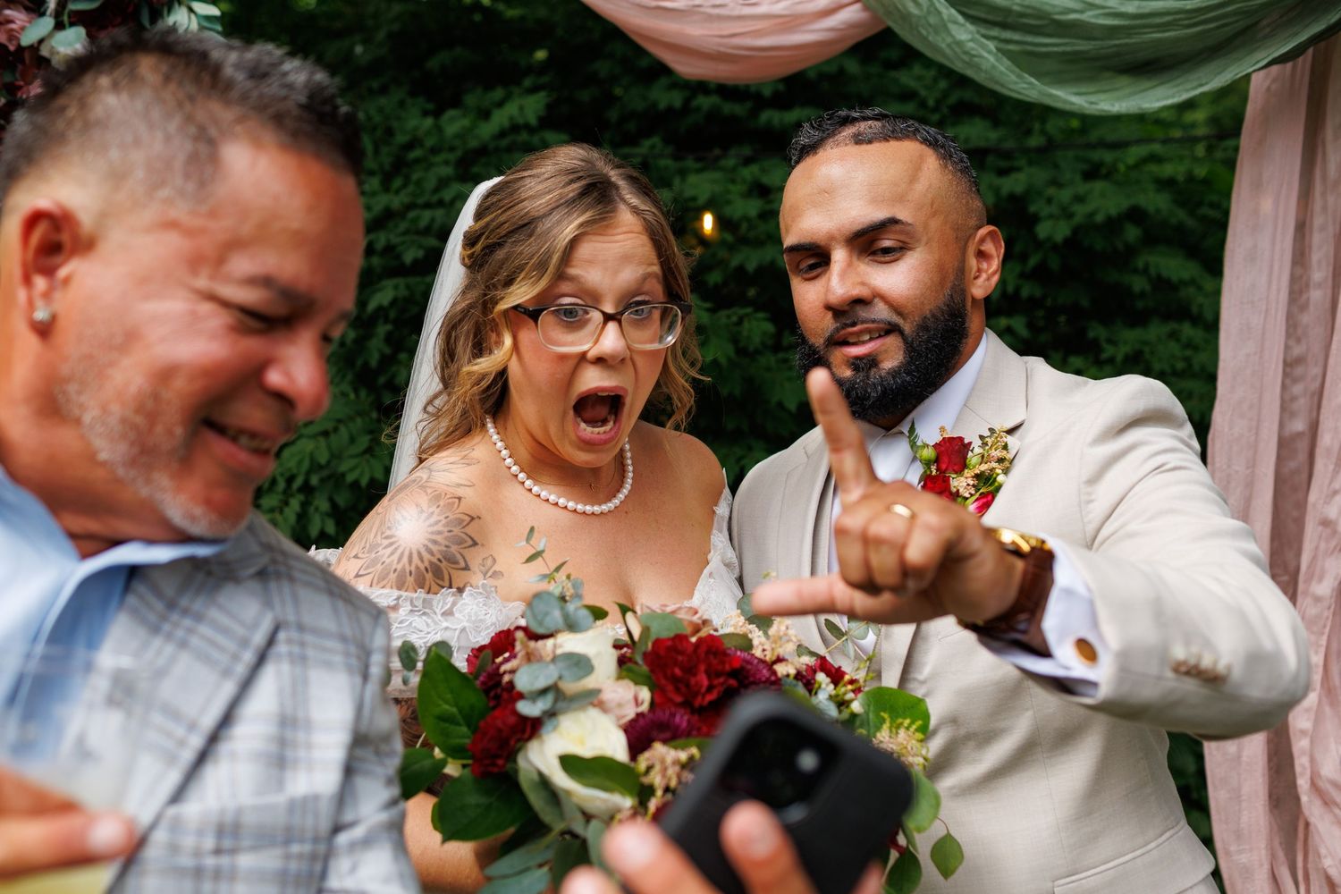 Bride and groom taking a selfie with a guest during outdoor wedding ceremony.