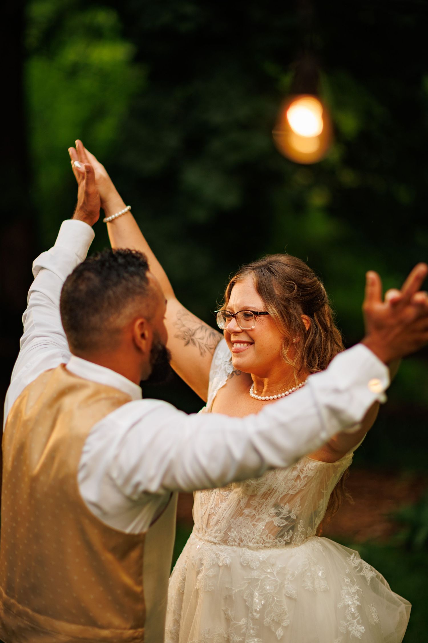 Bride and groom share a joyful first dance under warm string lights surrounded by lush greenery.