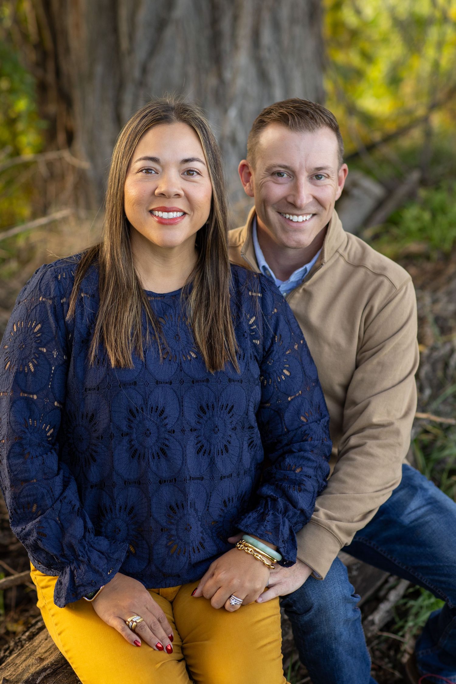 A couple sits together outdoors against a backdrop of autumn foliage and sunlit trees.