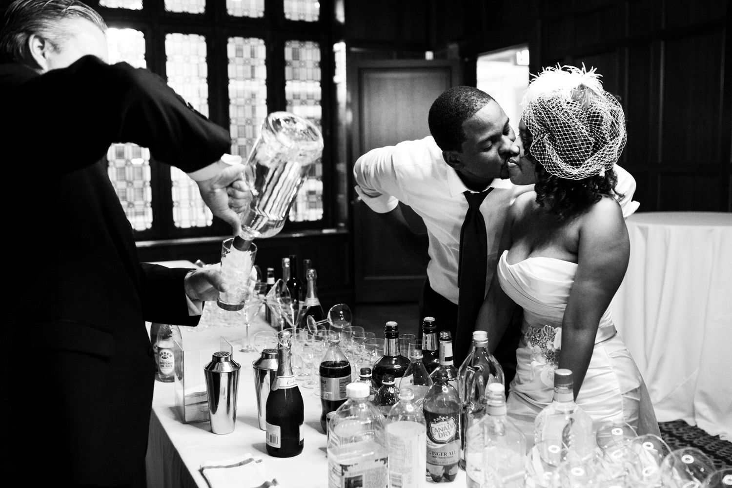 a black and white photo of a wedding couple kissing at the bar while the bartender ppours a drink for them