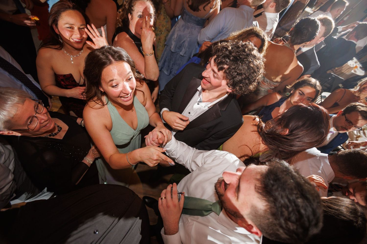 Excited wedding guests laugh and dance together in a lively crowd at an outdoor evening reception.