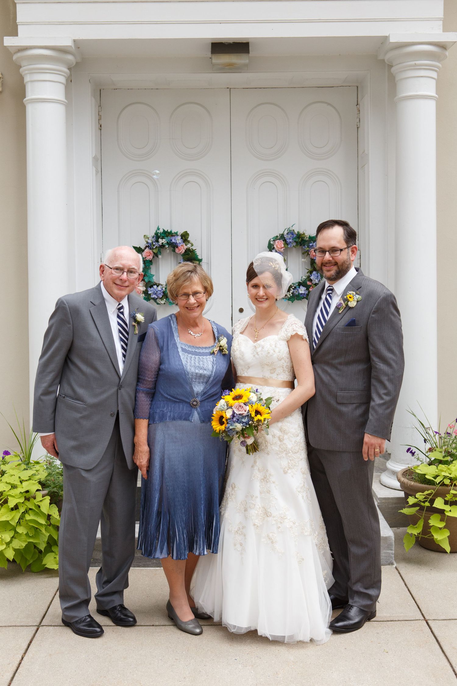 A formal wedding party poses together in front of white columns and door decorated with floral wreaths.