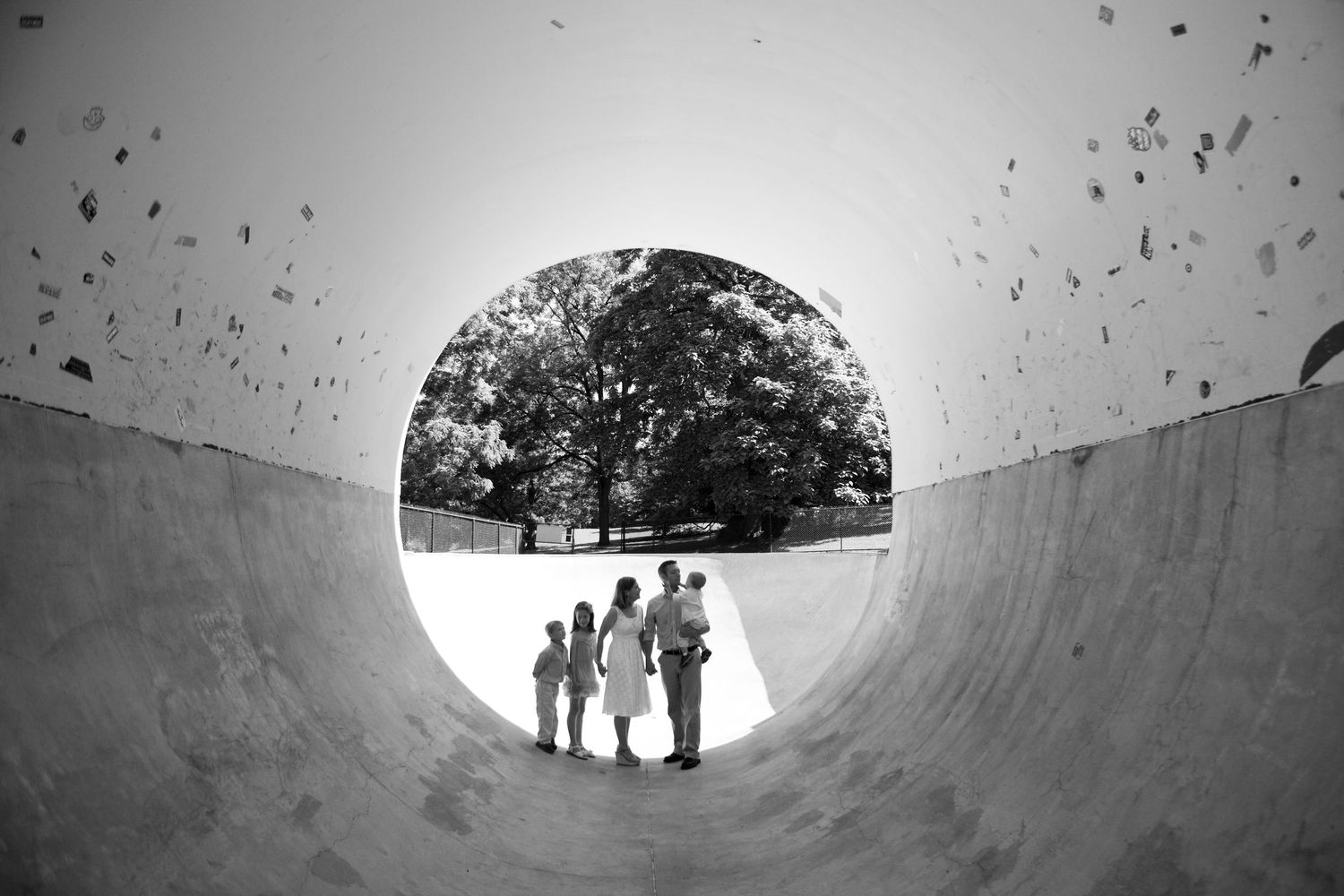 Black and white view through circular skate ramp showing silhouettes of people standing in front of trees.