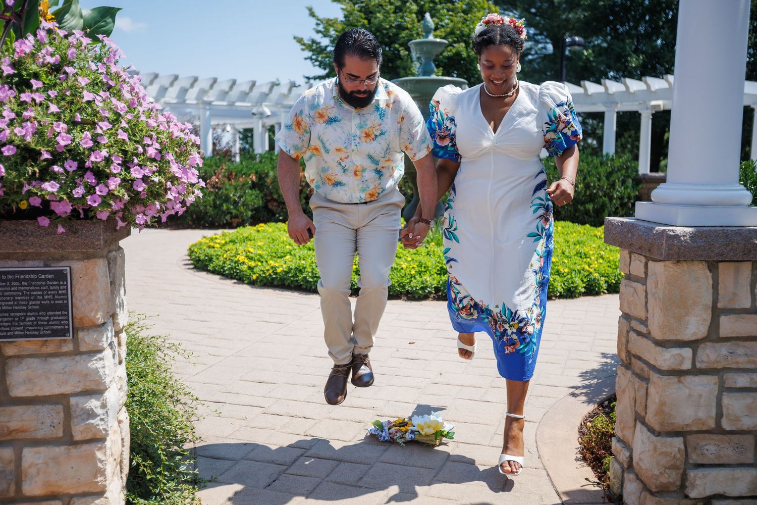 Couple holding hands walking through a beautiful garden with colorful flowers and white pergola on a sunny day.