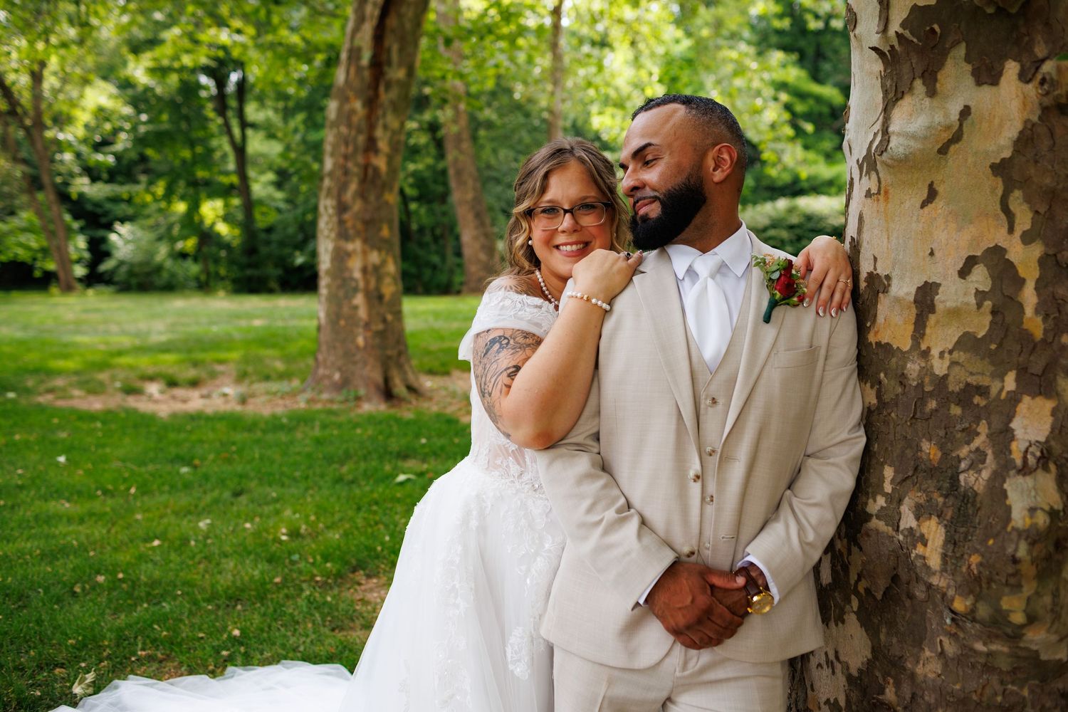 Bride and groom share a romantic moment leaning against a tree in a lush green garden setting.