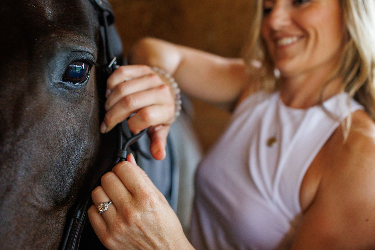 Woman smiling while adjusting a dark horse's bridle up close in a barn.