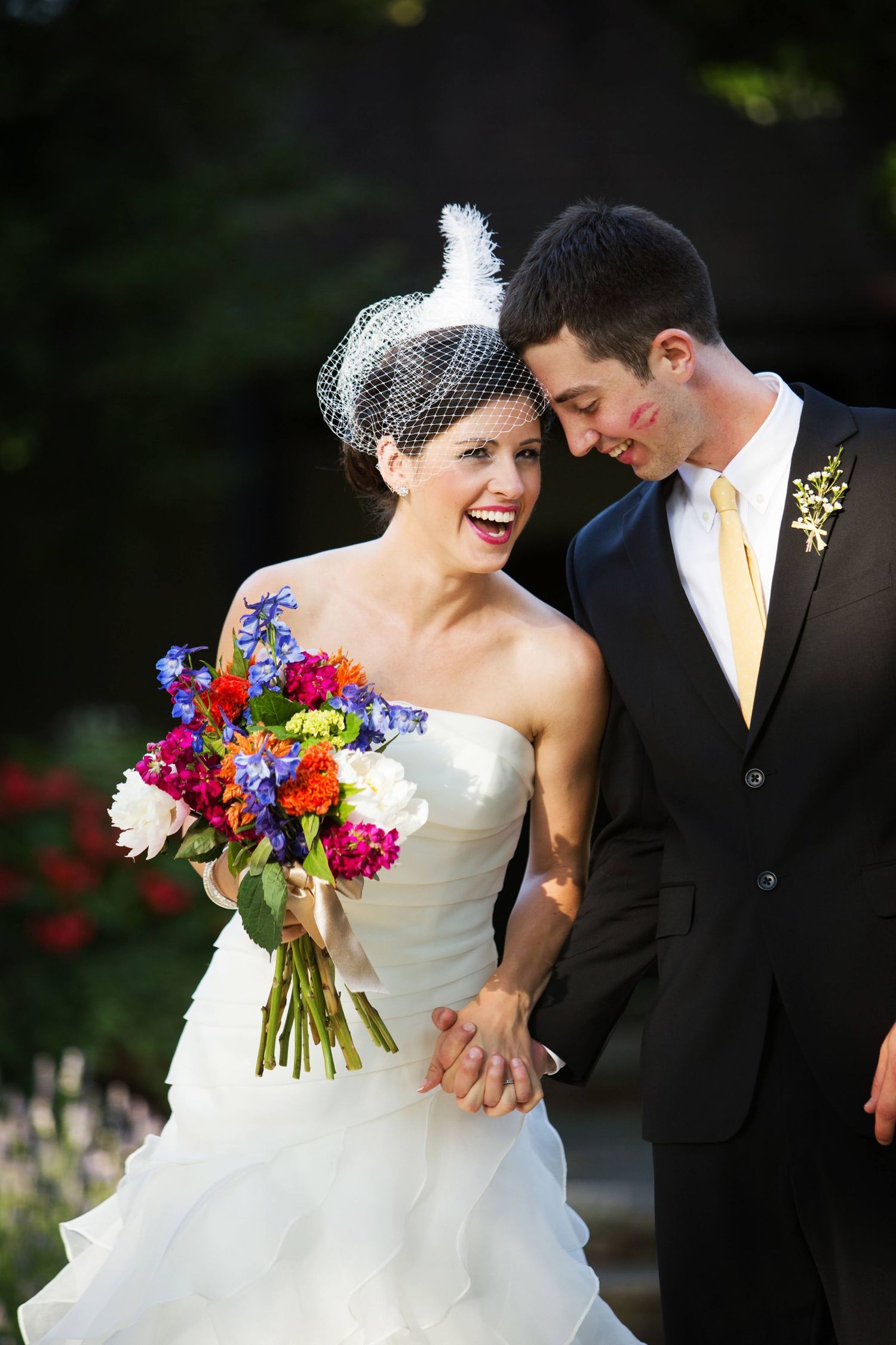 A bride in a white gown and birdcage veil holds a vibrant bouquet of red, blue and pink flowers next to a groom in black suit.