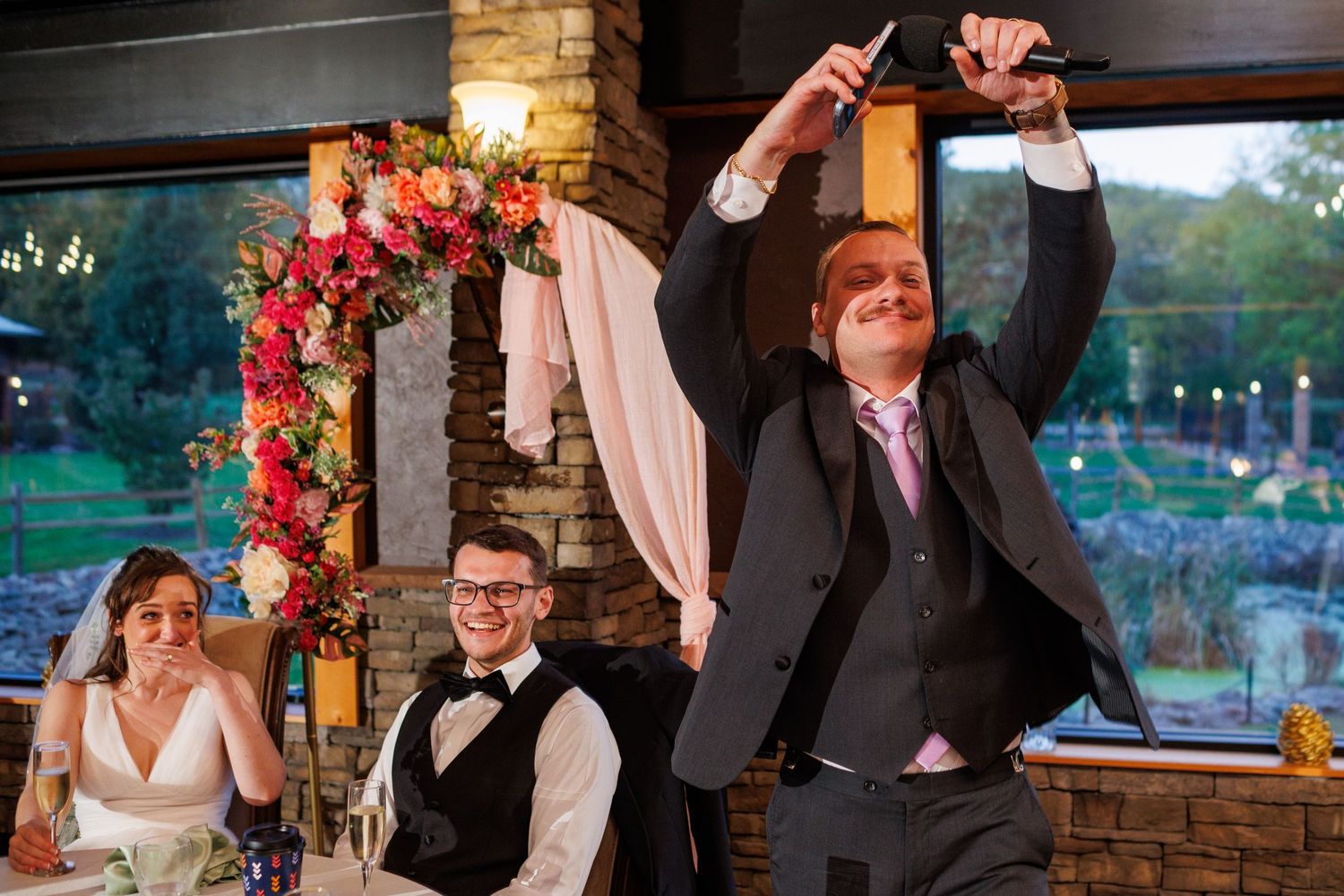 Red-haired groomsman raises knife while giving toast at colorful floral wedding reception.