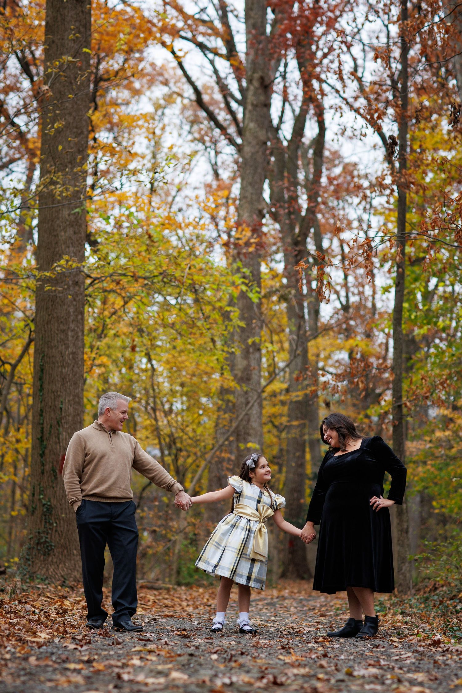 Three people hold hands while walking along a leaf-covered forest path surrounded by autumn trees with golden foliage.