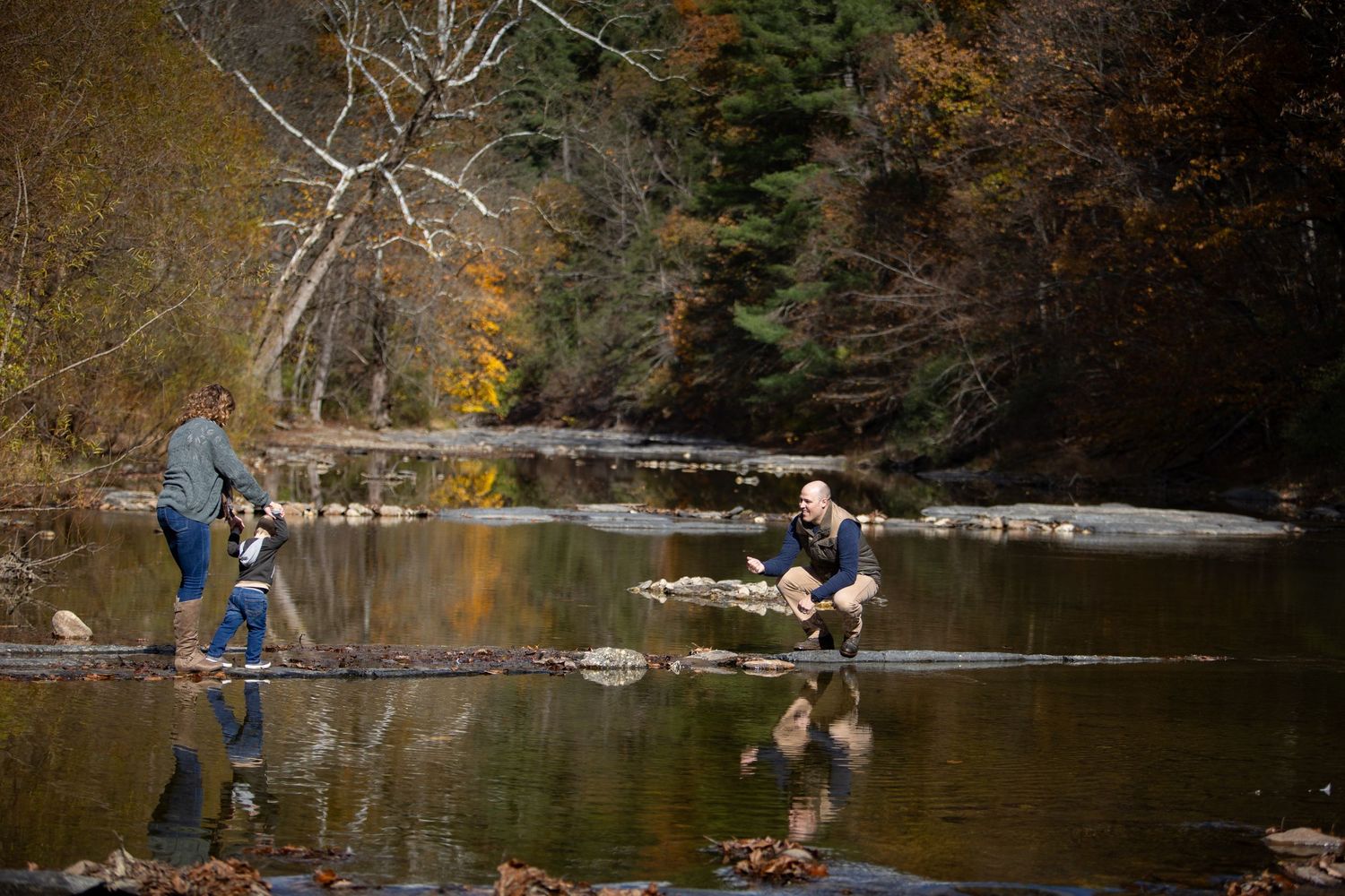 Two people fishing in a scenic autumn stream with fallen logs and colorful foliage reflecting in the calm water.