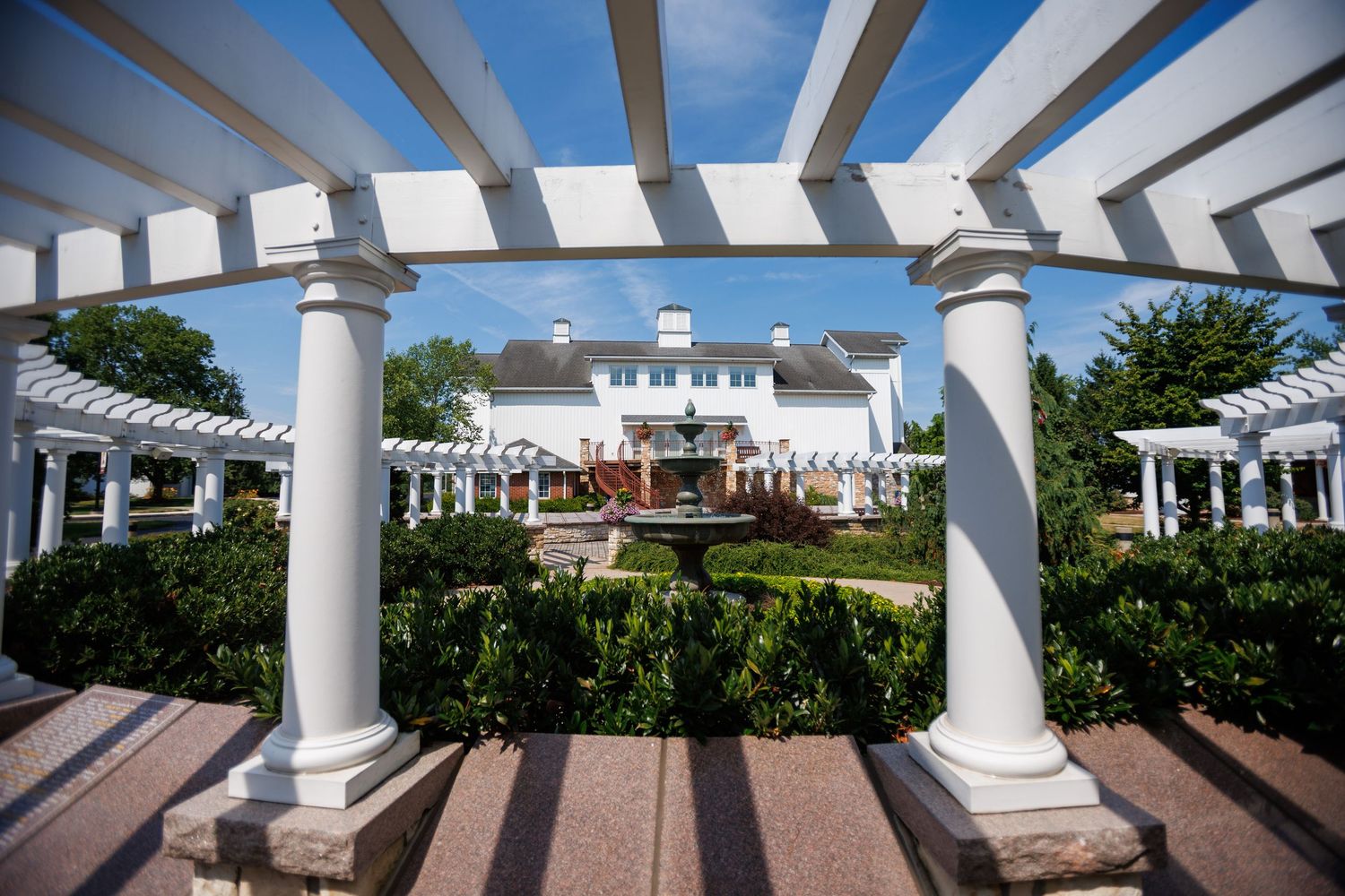 White pergola columns frame a elegant estate garden with fountain and manicured hedges under a blue sky.