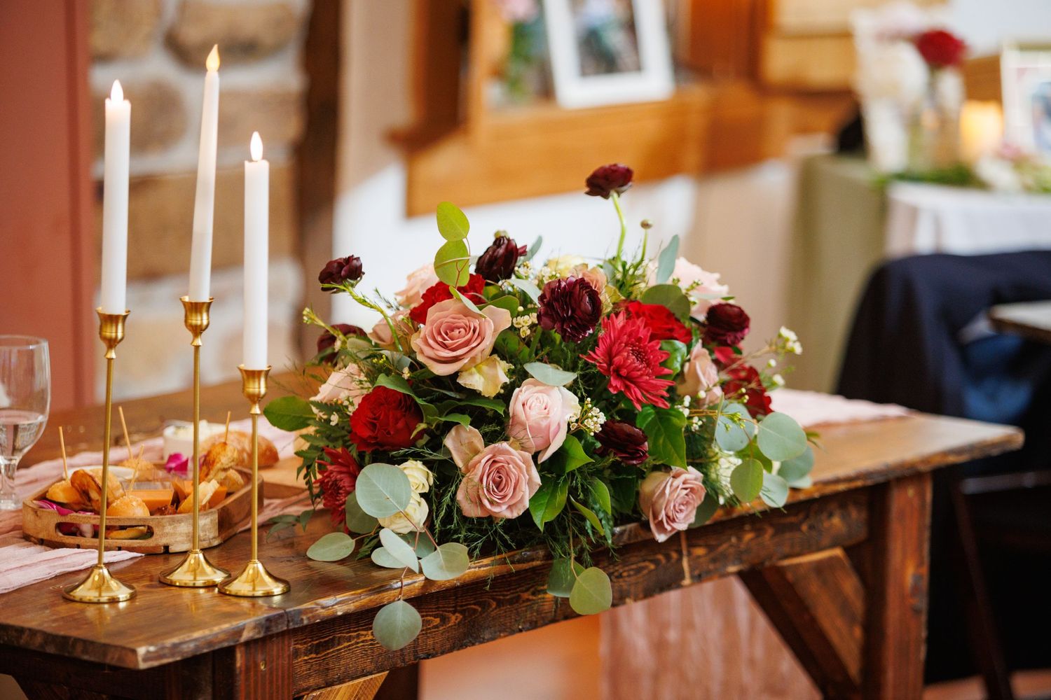 Lush floral centerpiece of red, pink, and burgundy roses with greenery beside gold taper candles on wood table.