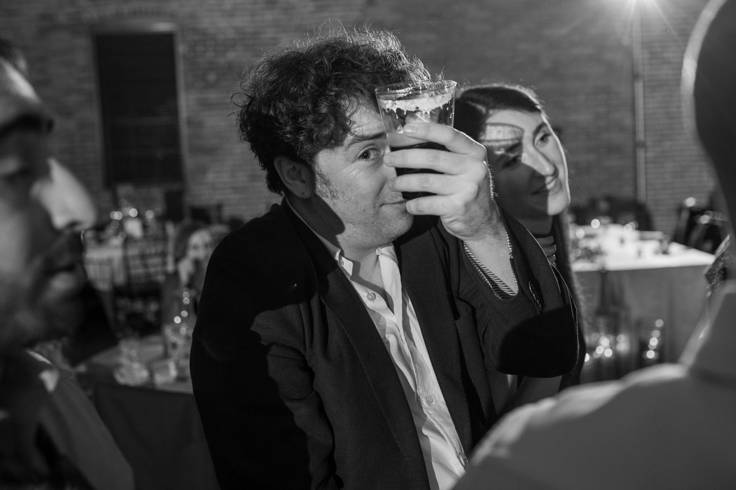 Black and white candid of wedding guest drinking from glass at lively evening reception celebration.