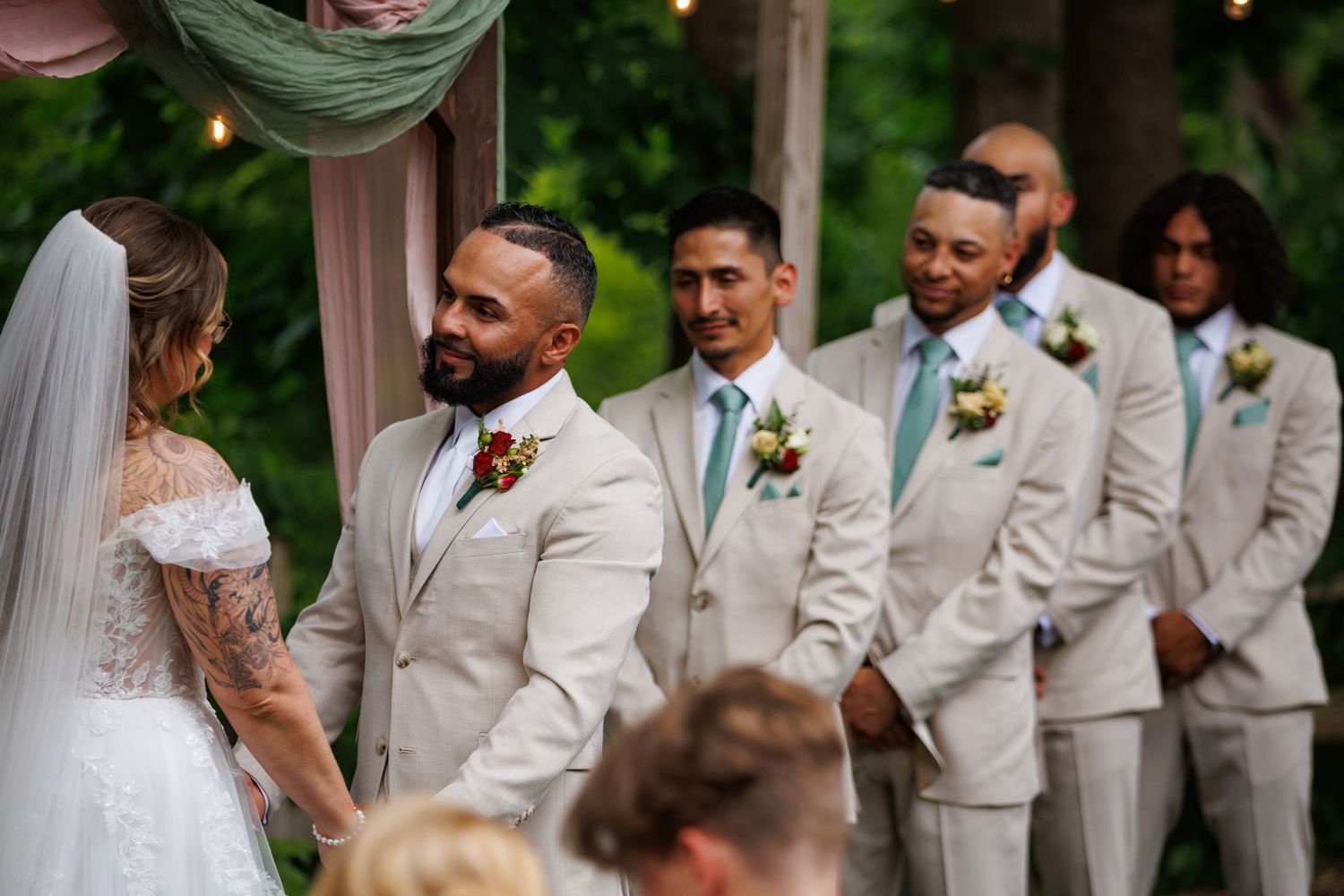 Groom in tan suit smiling at bride during outdoor wedding ceremony with groomsmen standing behind.