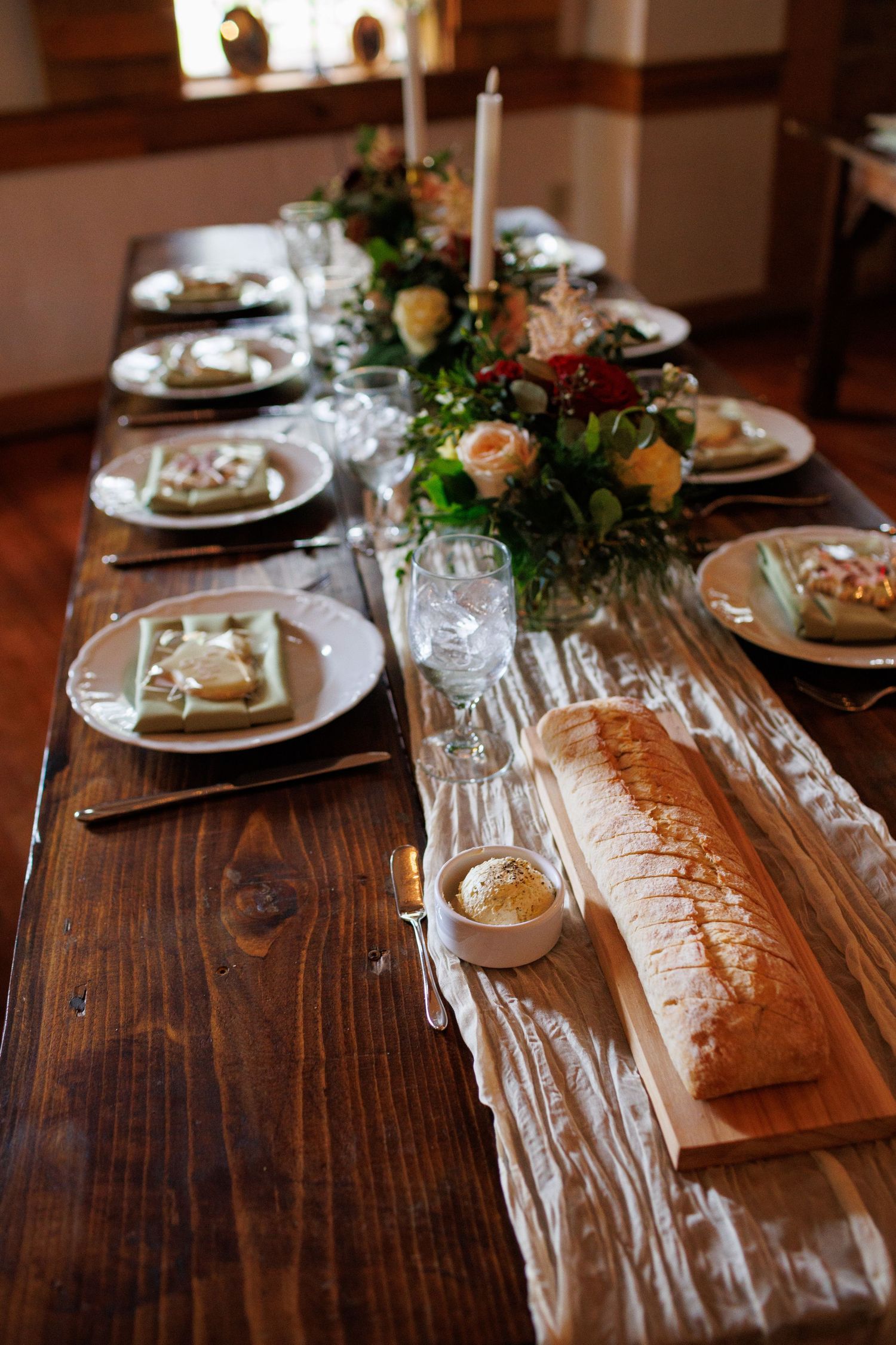 Rustic wooden farm table set with white plates, candles, floral centerpieces, and a sliced baguette on a cutting board.