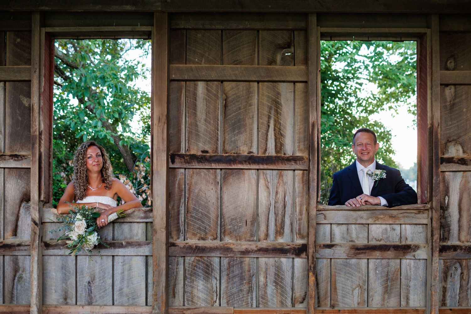 Bride and groom posing in separate windows of a rustic wooden barn on wedding day.