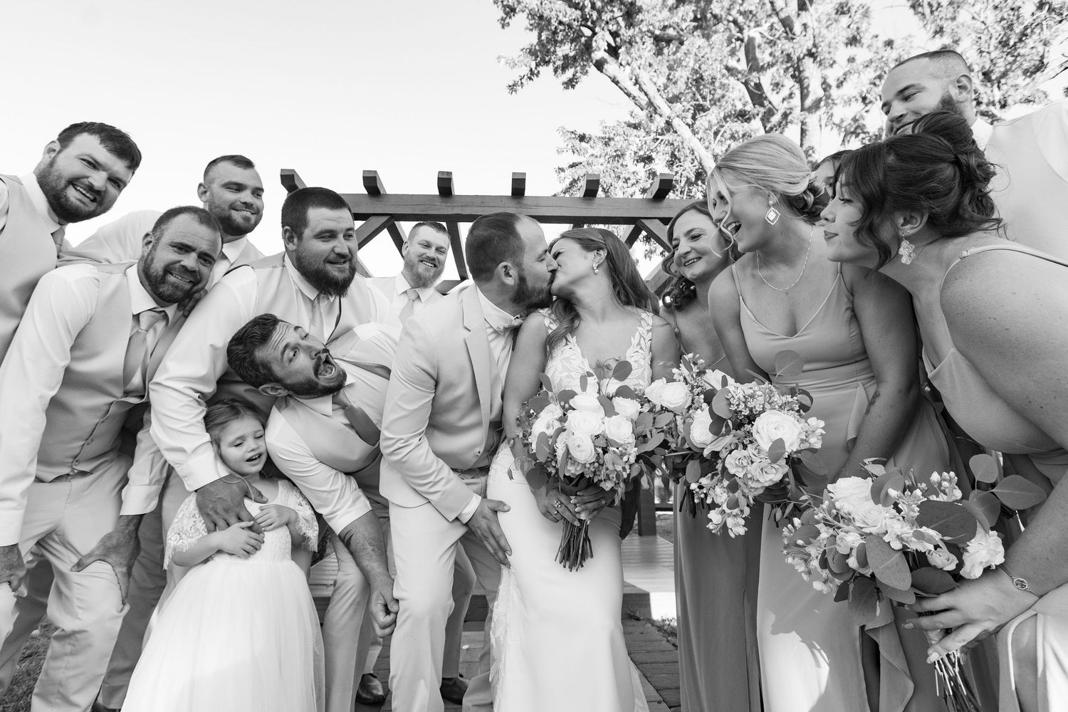 Black and white wedding photo of a bridal party gathered together sharing laughter and smiles under an outdoor arch.