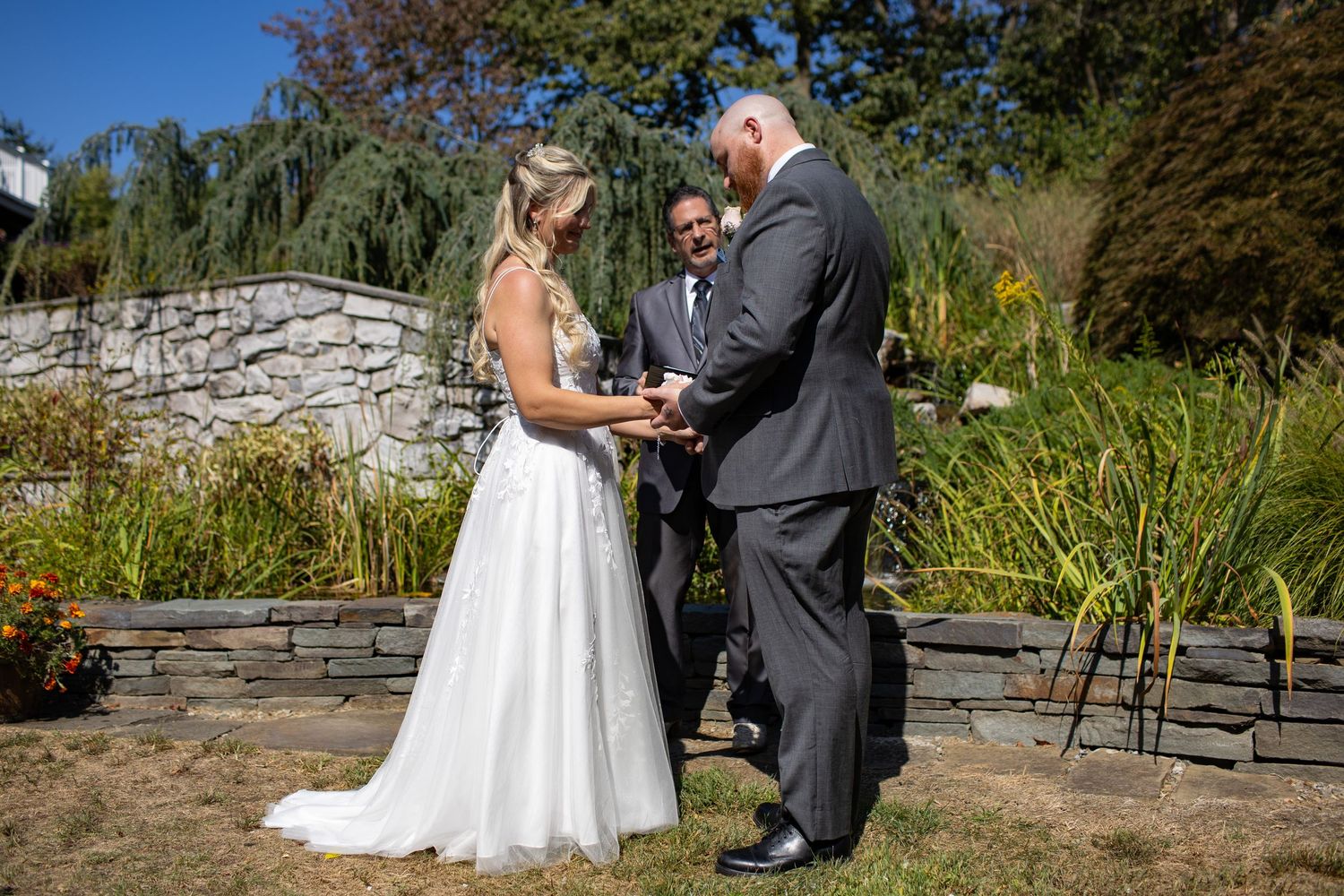 A couple exchanges vows during an outdoor garden wedding ceremony with stone walls and natural landscape in the background.